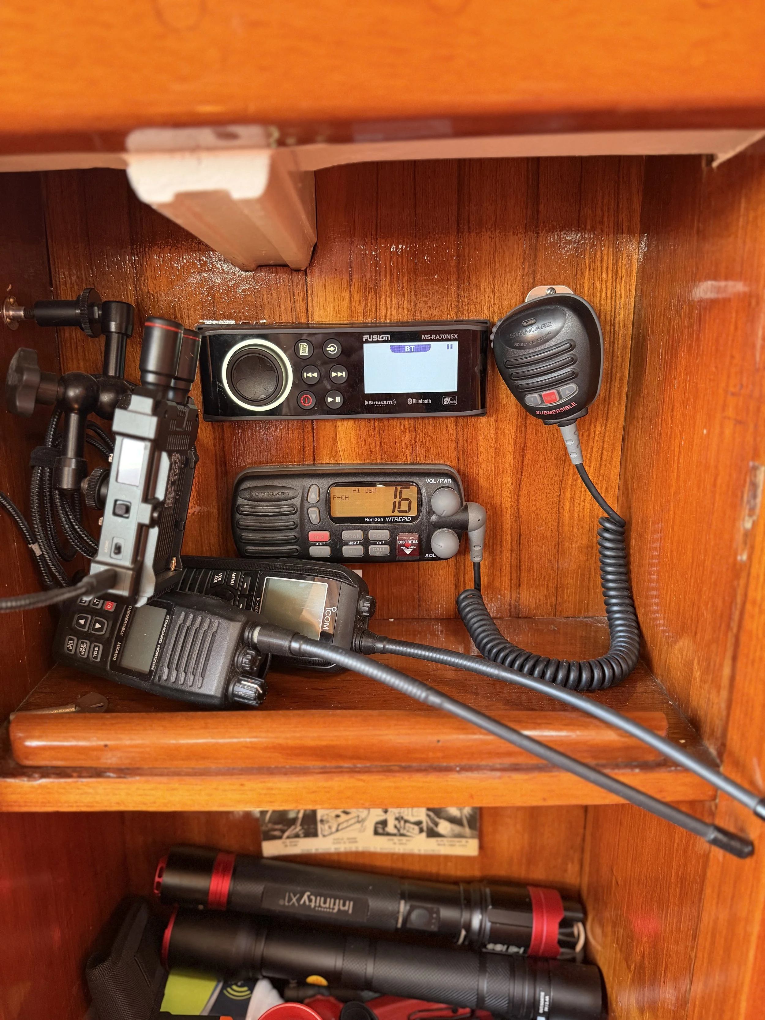 Close-up view of marine communication devices including a handheld radio, an amplifier, and a VHF radio, all stored in a wooden compartment.