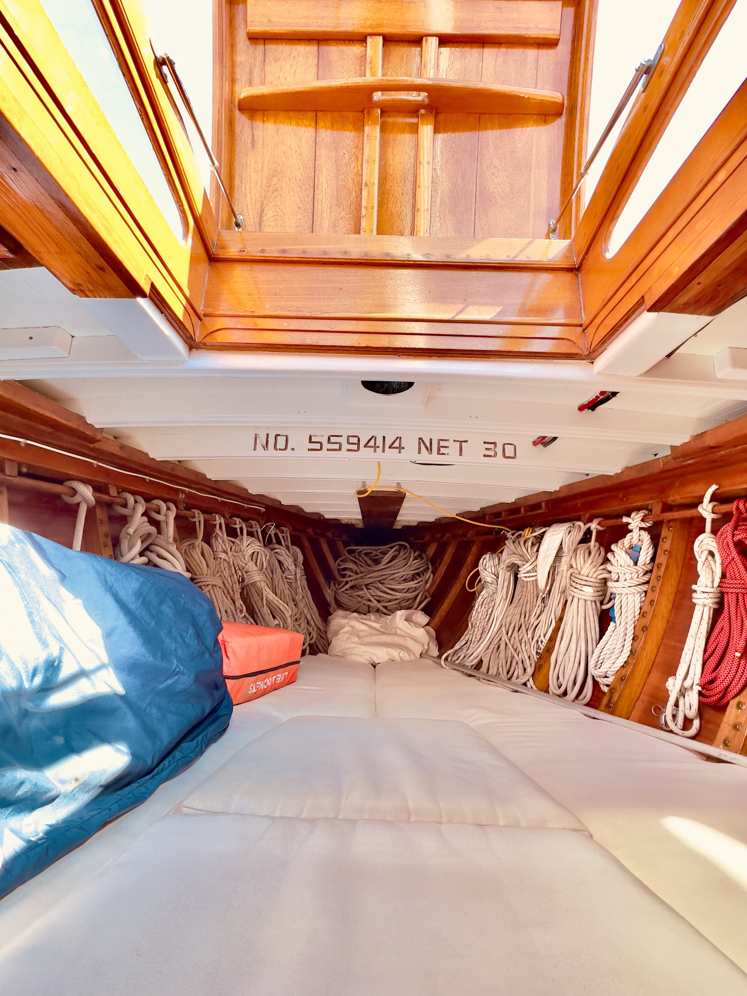 View inside a wooden sailing boat's cabin showing coiled and hanging ropes, a blue bag on the left, a red box, and the floor with a sleeping mat. The ceiling has a registration number 'NO. 559414 NET 30'.