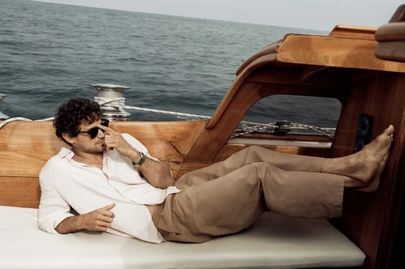 A man relaxing on a wooden boat, reclining on a cushioned surface with his feet up, wearing sunglasses and a white shirt, with ocean in the background.