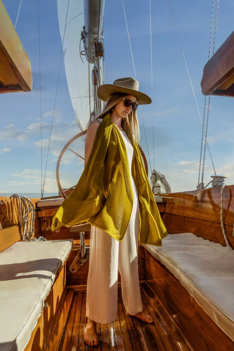 A woman wearing sunglasses, a wide-brimmed hat, a white dress, and a yellow shawl standing on a wooden boat deck under a clear blue sky.