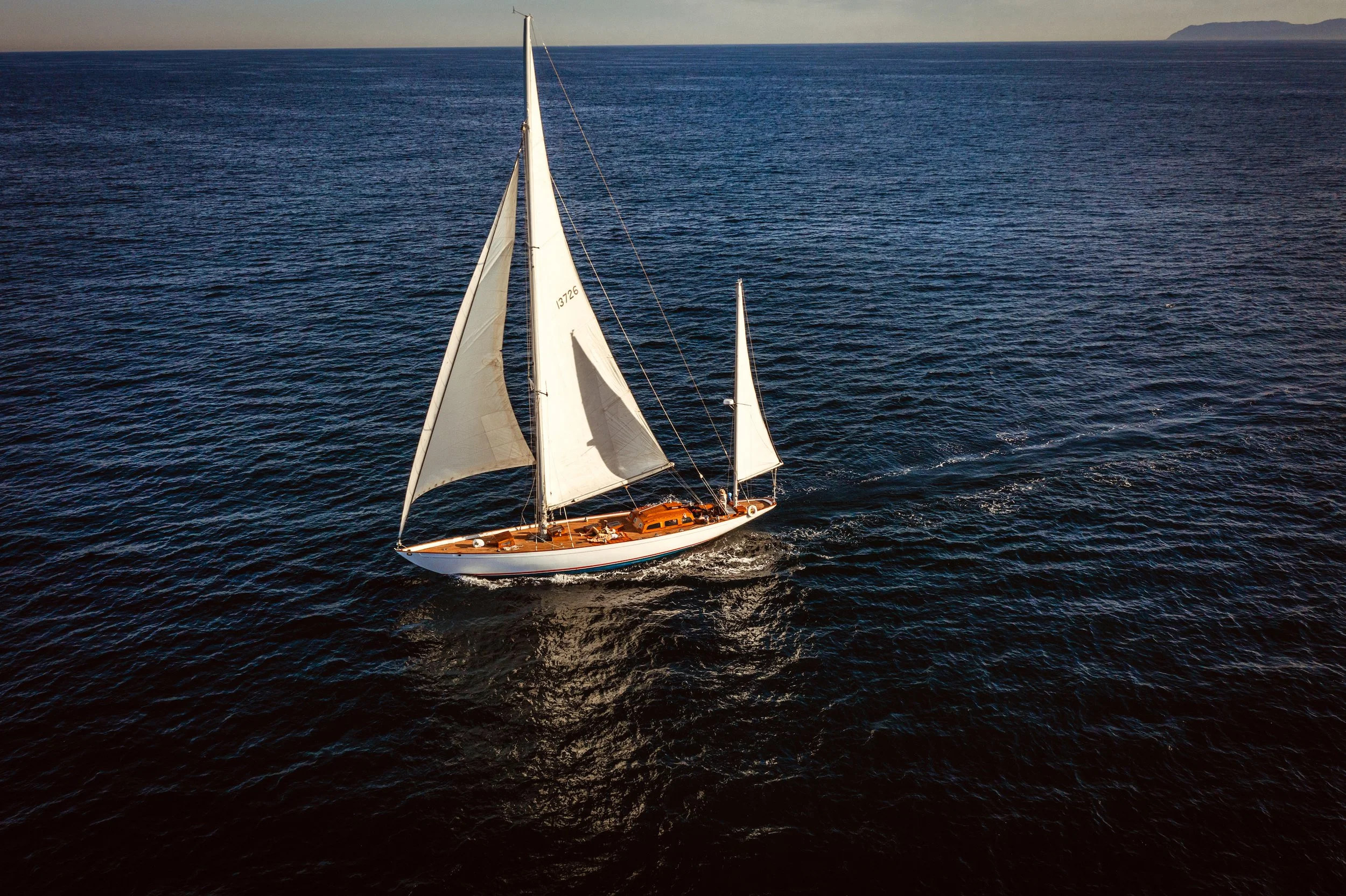 A sailboat with white sails and a wooden hull sailing on the ocean with a distant landmass on the horizon.