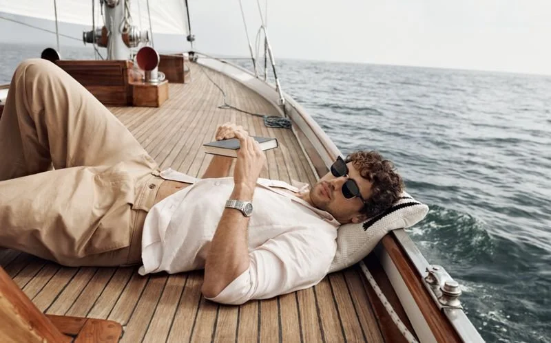 In the style of JFK Love Story, A man lying on a boat deck, wearing sunglasses and a white Calvin Klein shirt, looking at his phone.