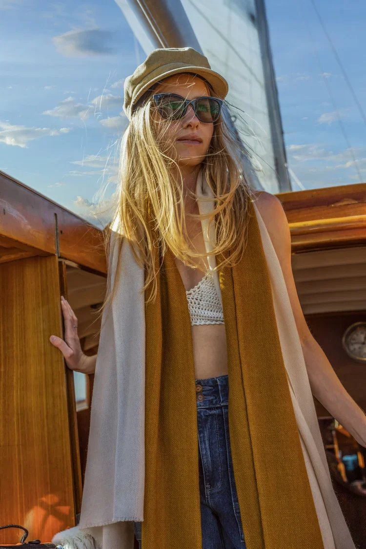 A woman with long blonde hair wearing sunglasses, a cashmere beige cap, a white crochet top, high-waisted jeans, and a long cashmere scarf, standing on a boat with a blue sky and clouds in the background.