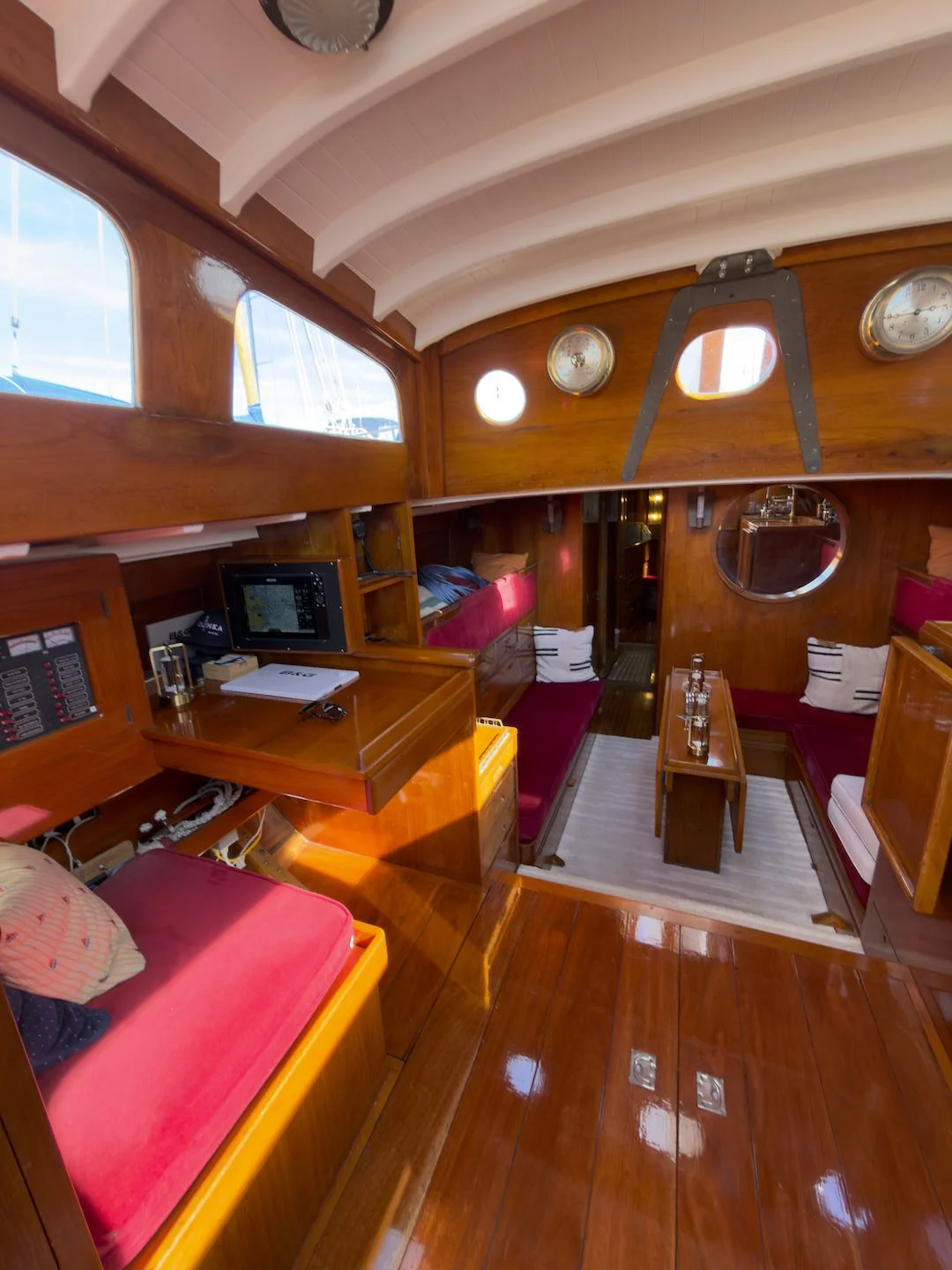 Interior of a wooden boat cabin with seating areas, a small table, and windows. The cabin features polished wood surfaces, red and white cushions, and nautical gauges on the wall.