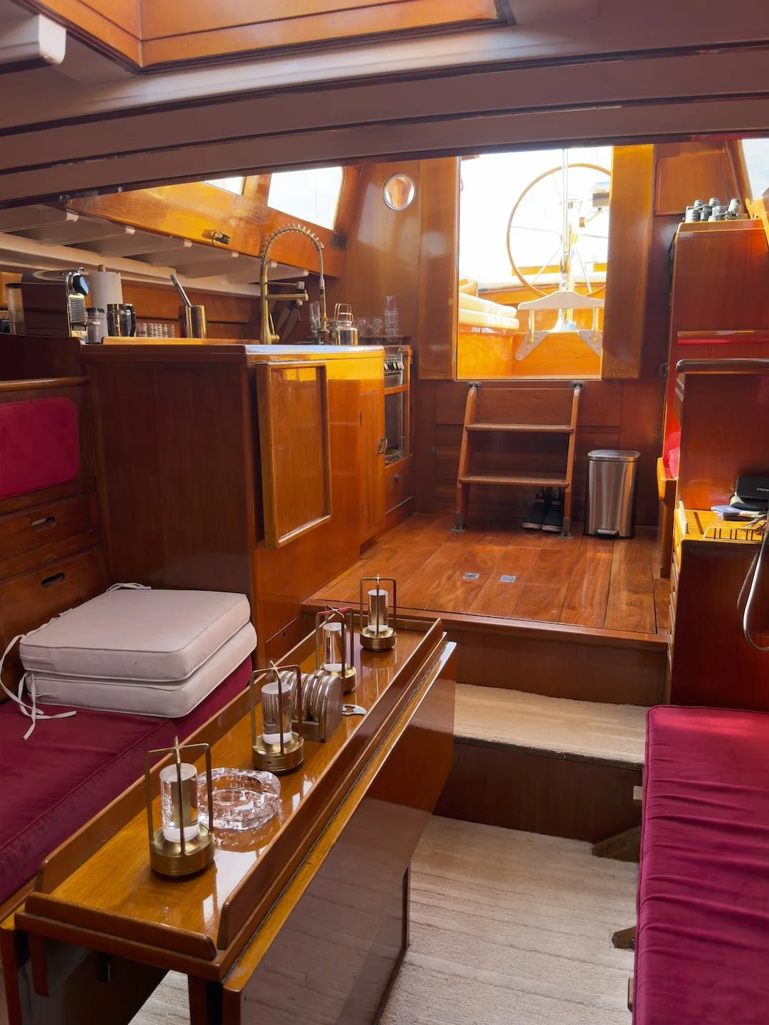 Interior of a boat cabin with wooden paneling, a small kitchen area, and seating. The room features a window with sunlight, a sofa with red cushions, a wooden table with decorative items, and various kitchen appliances.