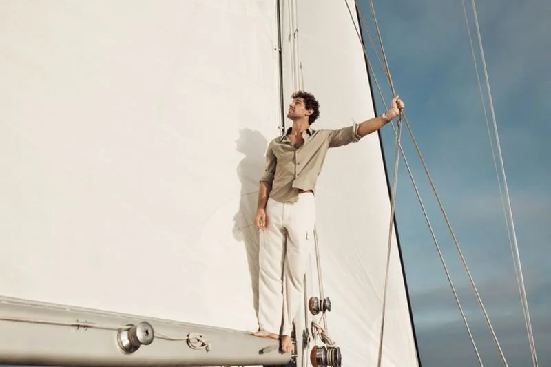 A man with curly hair and a beard standing barefoot on the edge of a sailboat with a large white sail, holding onto the rigging, looking up with a relaxed expression during daytime.