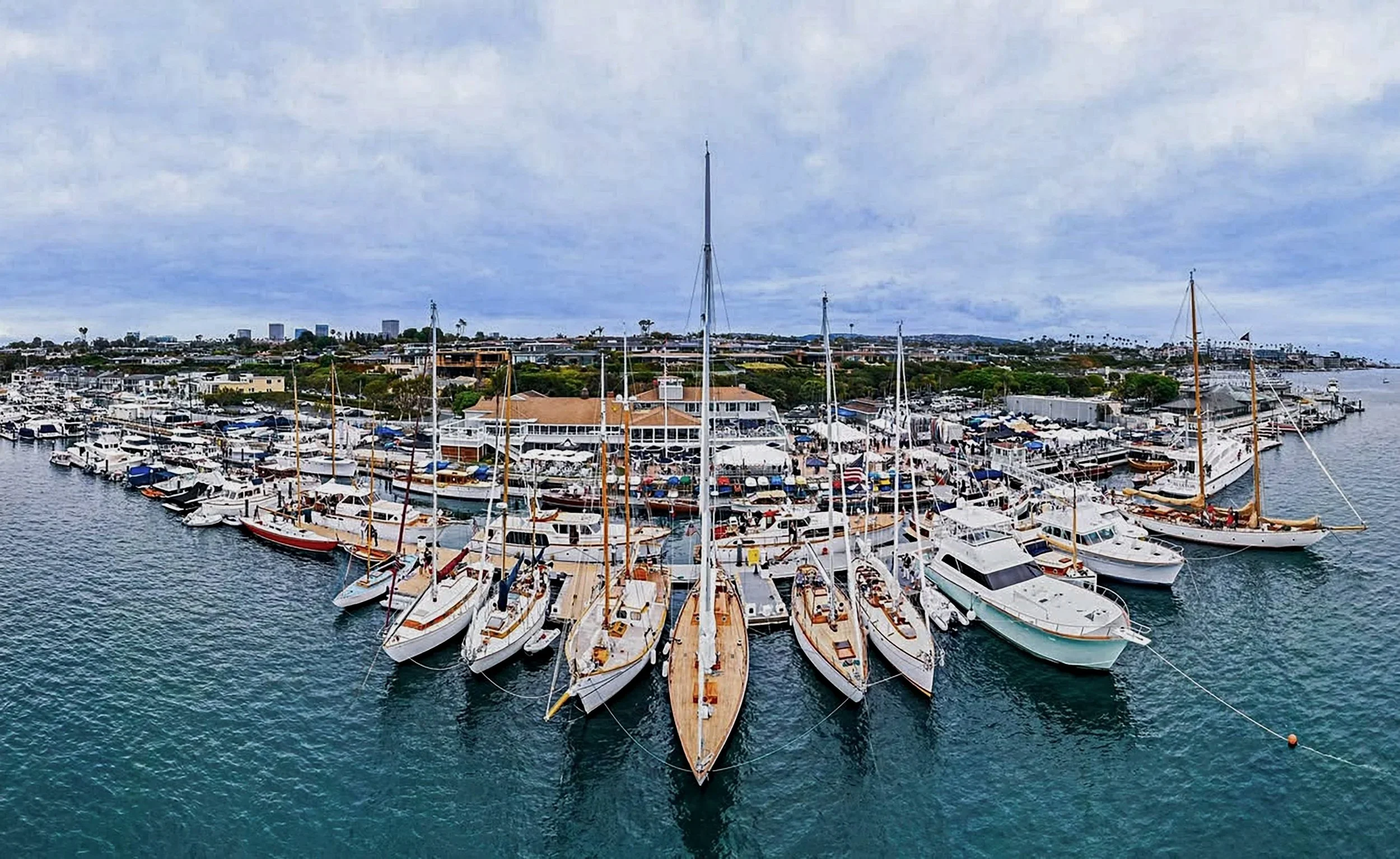Aerial view of a marina with numerous docked sailboats and yachts, a clubhouse with outdoor seating, and buildings in the distance under a cloudy sky.
