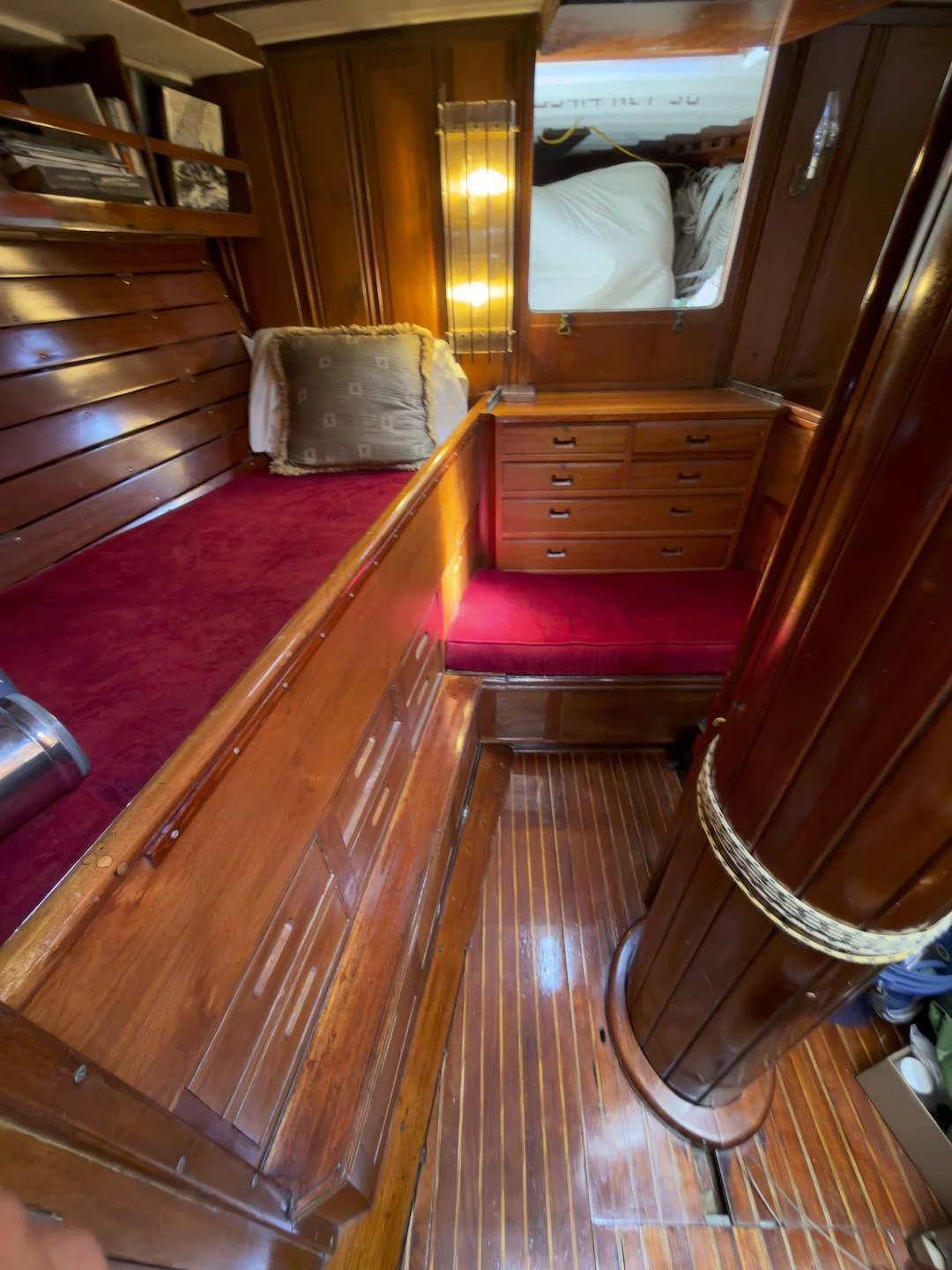 Interior of a boat cabin with wooden paneling, a narrow bed with a red mattress and a pillow, a window above, and a built-in dresser with multiple drawers.