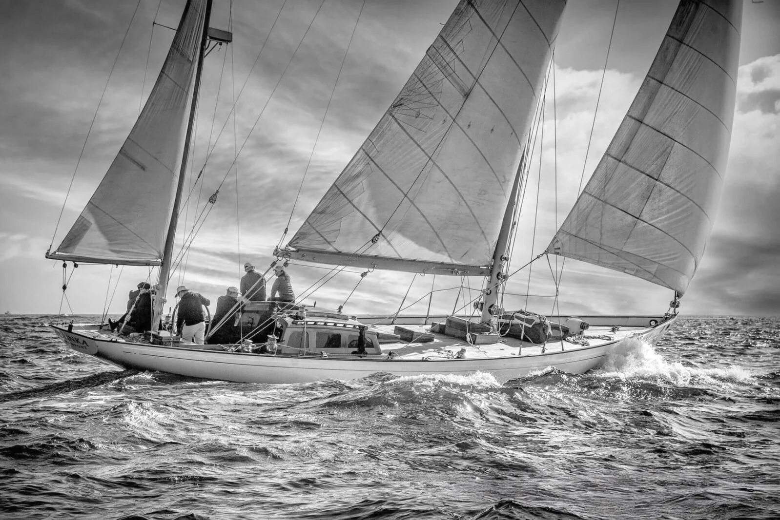 A black and white photo of a sailing yacht with multiple sails, crew members onboard, on choppy water under a cloudy sky.