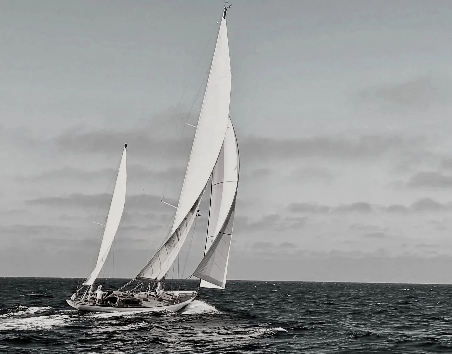 A sailboat with multiple large sails cruising on the ocean under a cloudy sky.