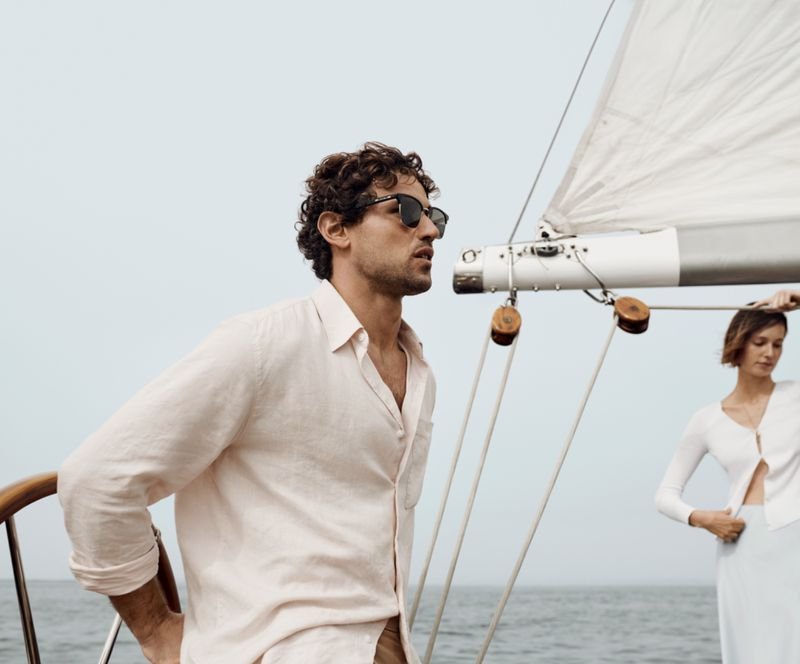 A man with curly hair wearing sunglasses and a white shirt on a sailboat, with a woman in a white dress in the background, and the ocean in the distance.