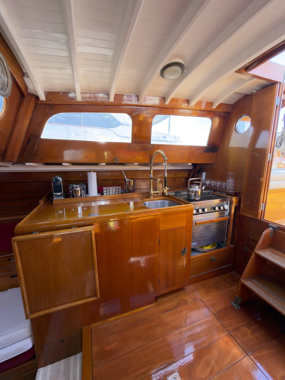 Inside a boat's kitchen with wood-paneled cabinets, a sink with a gold faucet, a stovetop, and overhead windows showing water outside.