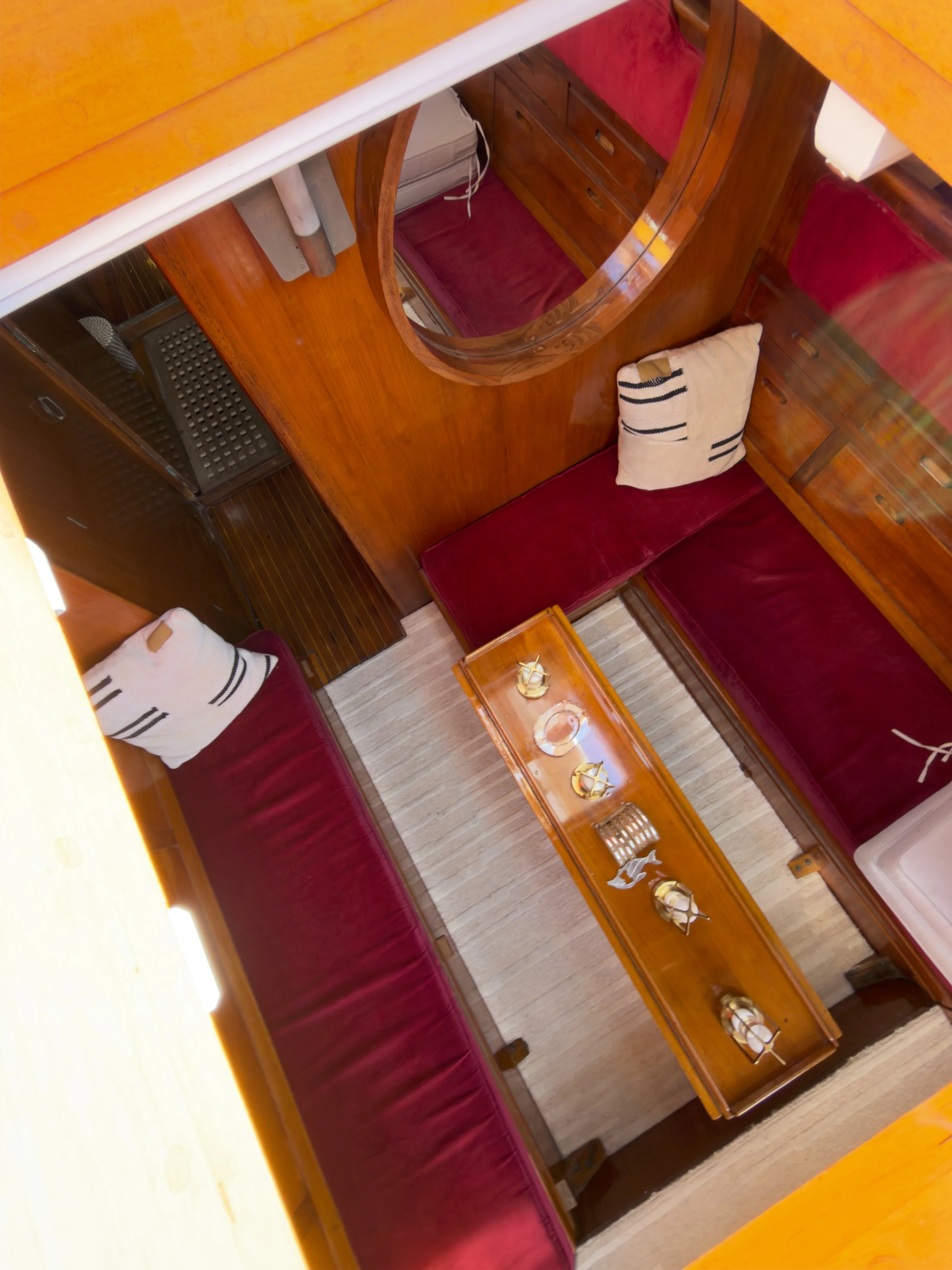 Inside a boat cabin with red cushioned seating, wooden walls, shelves with drawers, and a table with decorative items and cups on a striped rug.