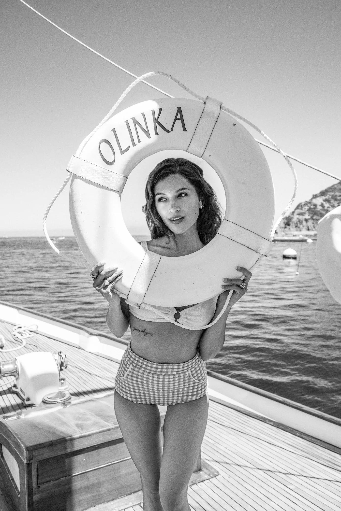 A woman in a bikini top and checkered shorts holds a life preserver with the word 'OLINKA' on it, standing on a boat near the water.