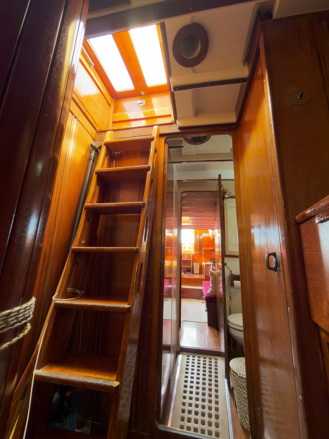 Interior of a boat with wooden walls, a ladder leading up to a skylight, a mirror, a doorway to a bathroom, and cozy furnishings visible through the doorway.