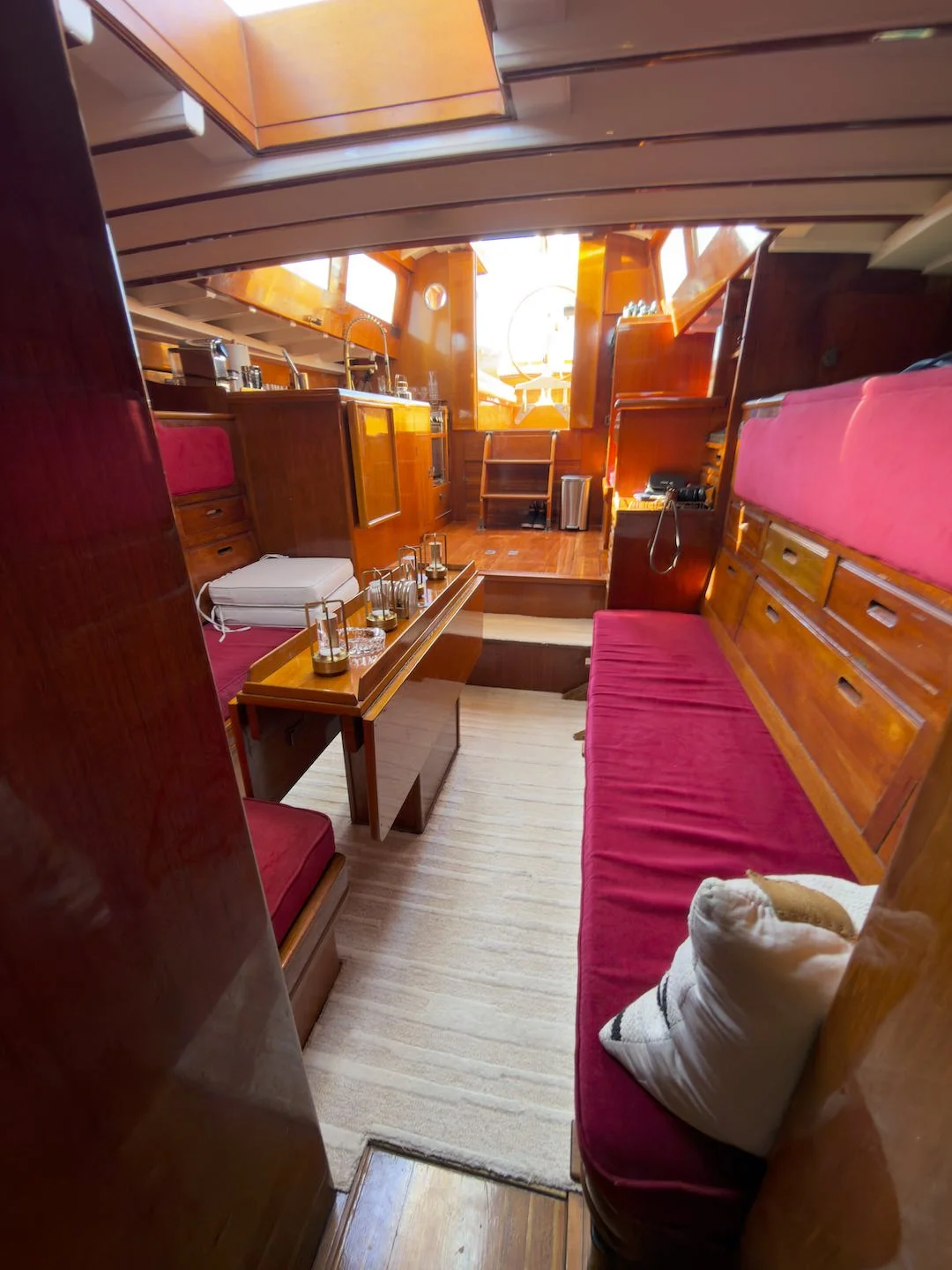 The interior of a boat or yacht with wooden paneling, pink cushioned seating, a small table with candles, and a view of the cockpit or outdoor area with a steering wheel in the background.