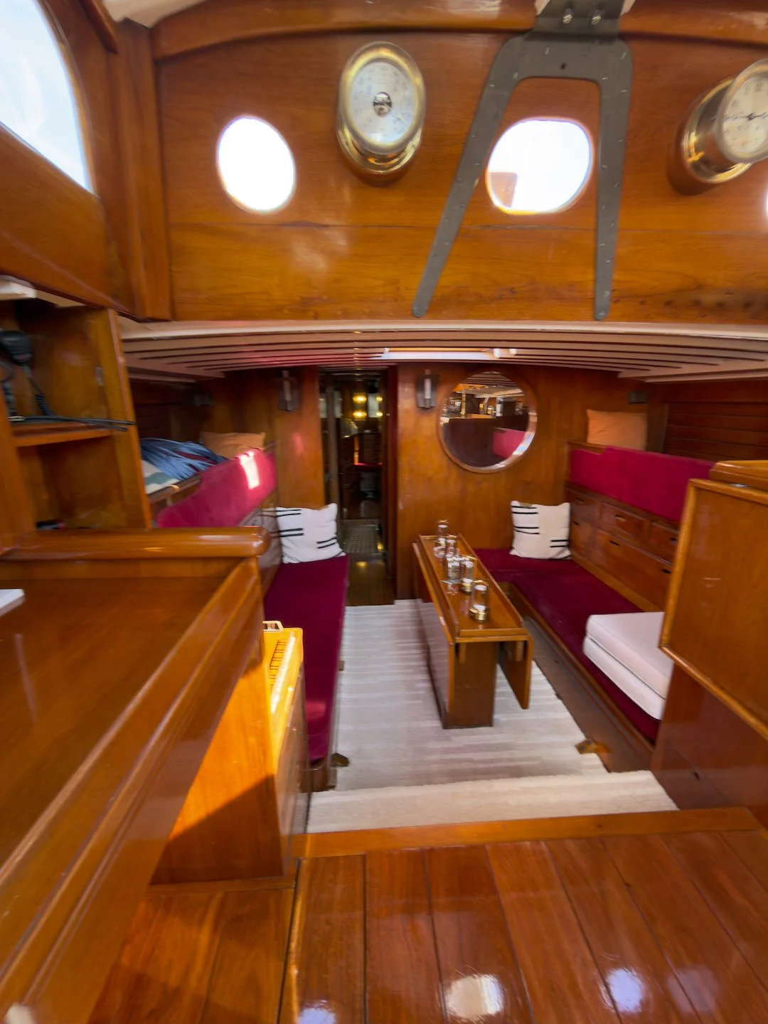 Interior of a wooden boat with red cushioned seating, a small wooden table set with glassware, round windows, and nautical instruments on the ceiling.