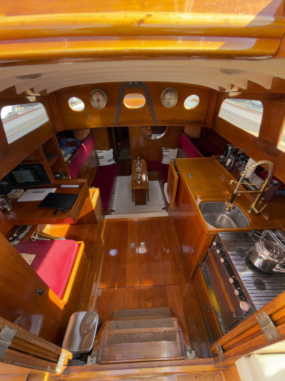 Interior view of a wooden boat cabin with a small kitchen area, a seating area with red cushions, and navigation dials on the wall.