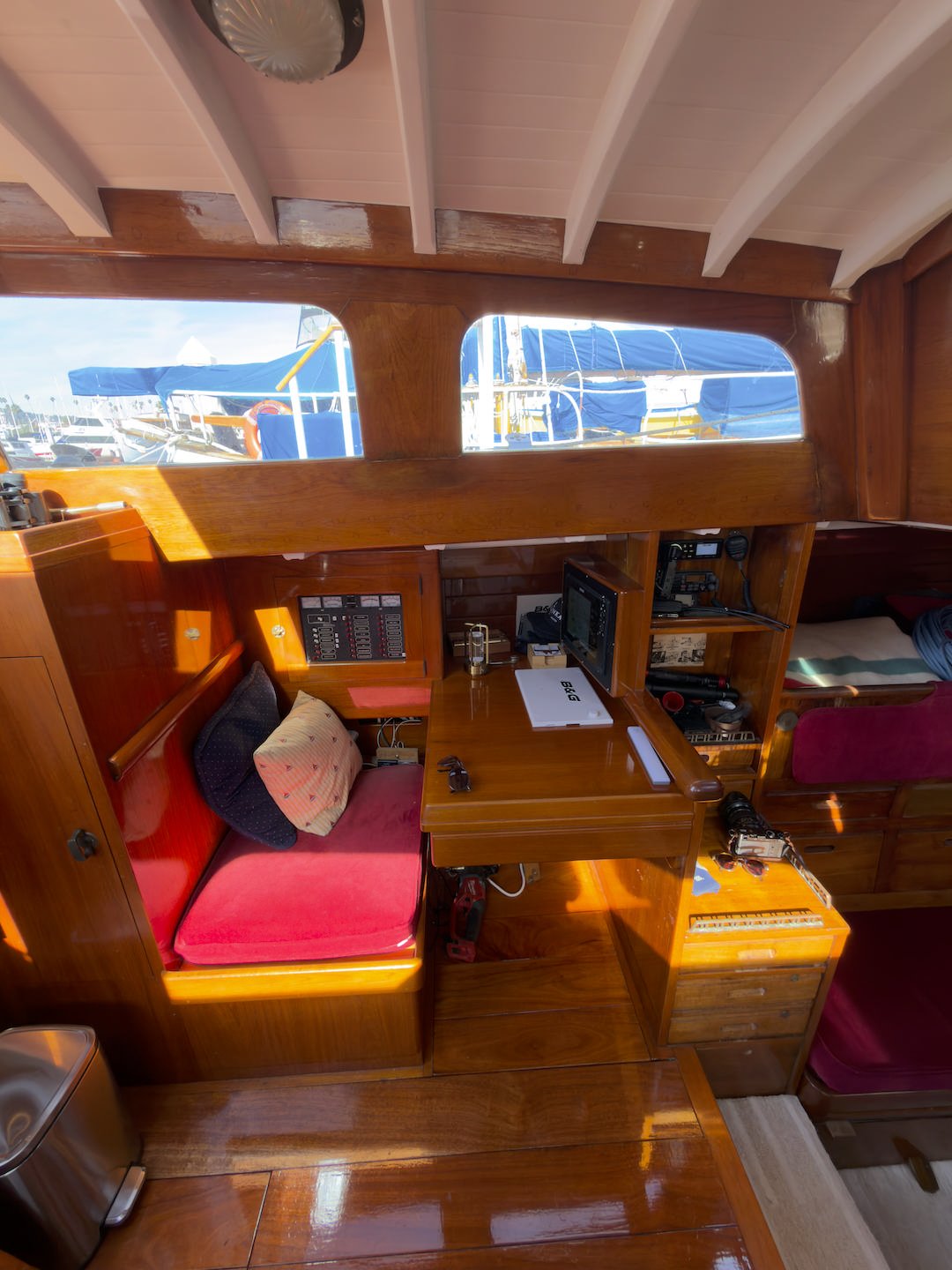 Interior of a boat cabin with wooden paneling, a small table, a cushioned bench with pillows, navigational and communication equipment, a computer monitor, and windows showing boats docked outside.