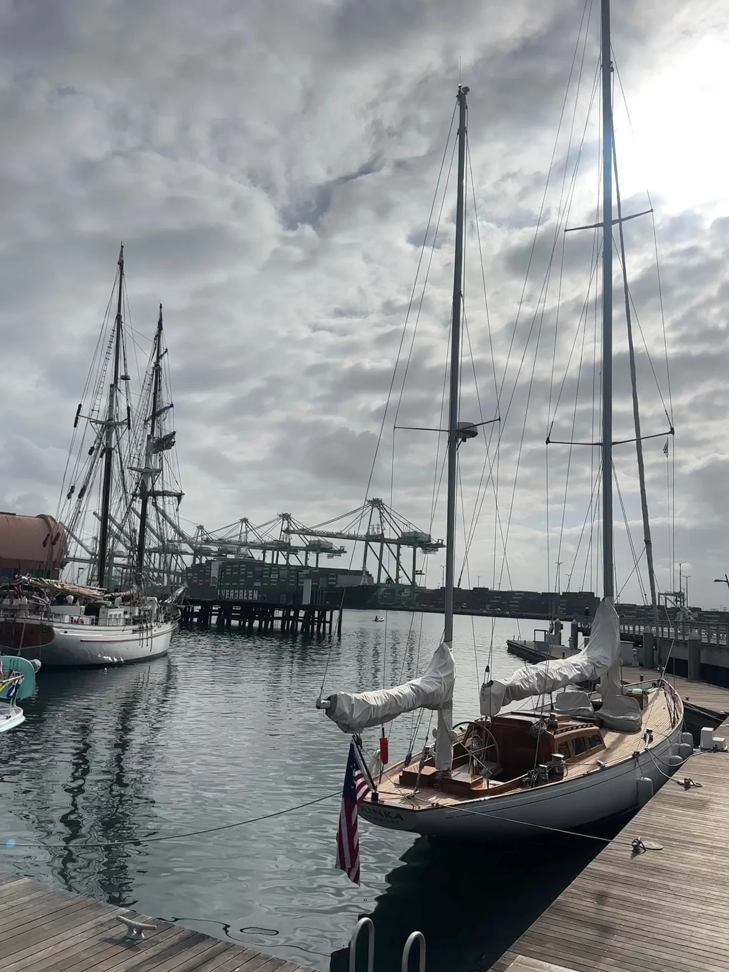 A sailboat docked at a marina with a wooden dock in the foreground, other boats, and a container port with cranes in the background, under a partly cloudy sky.