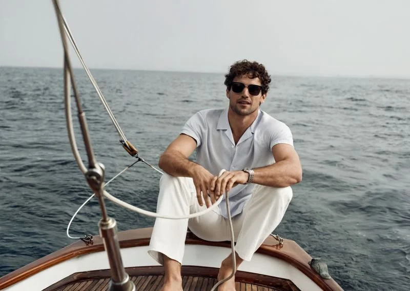 A man with curly hair and sunglasses sitting on the bow of a boat on the water.