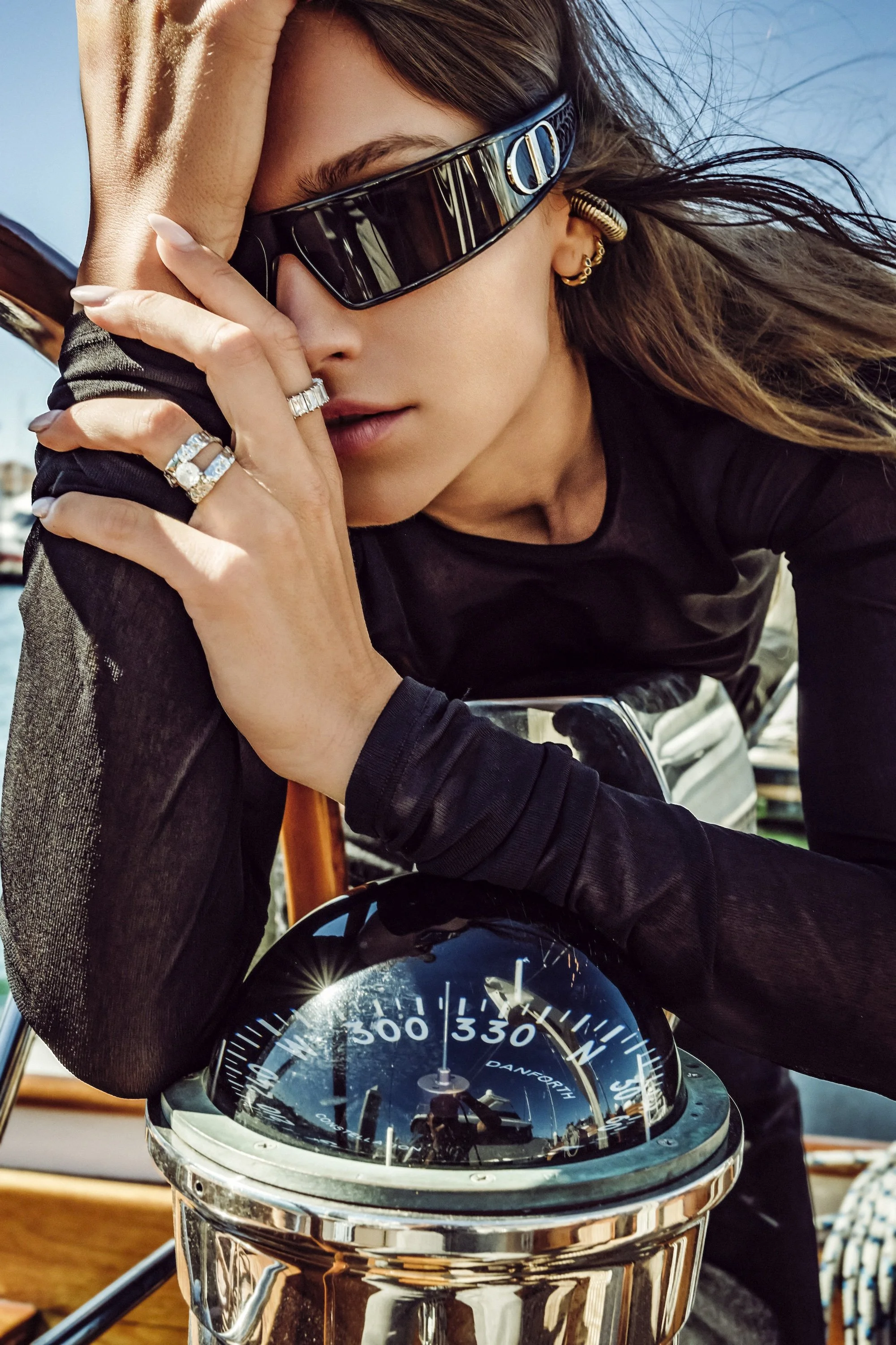 A woman with long hair wearing large black sunglasses, gold earrings, and rings on her fingers. She is leaning on a polished chrome nautical compass mounted on a wooden boat and looking at the camera.
