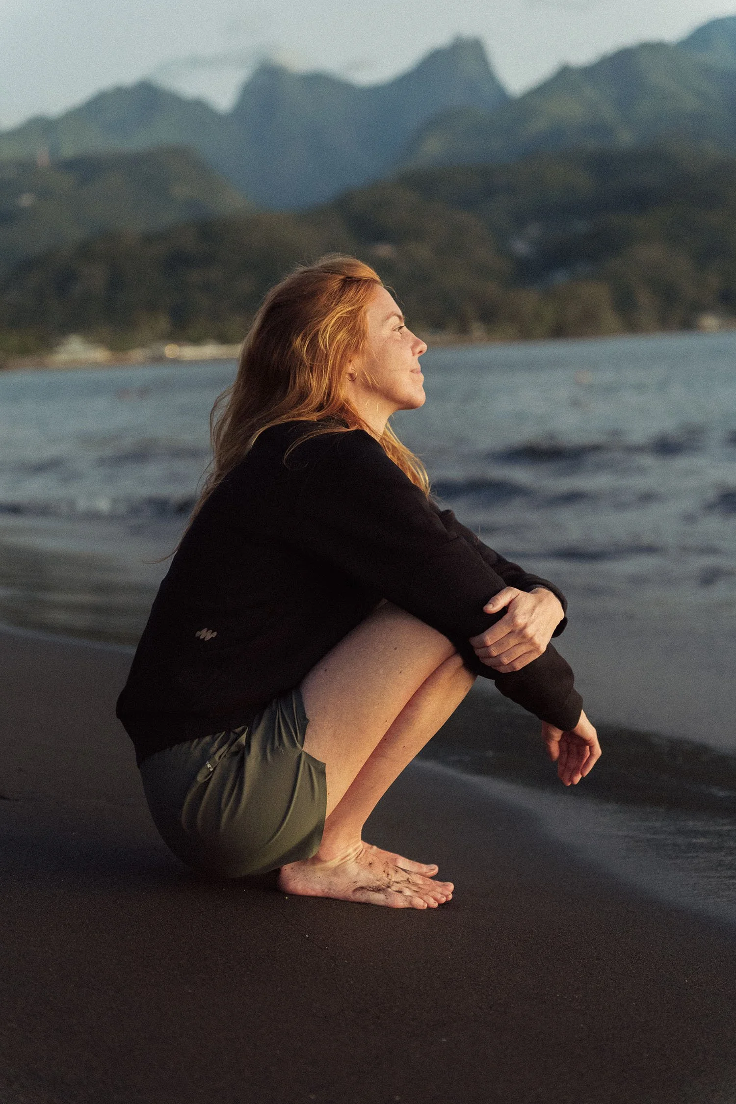 Karina Durand accroupie sur la plage, regardant vers la mer avec des montagnes en arrière-plan.