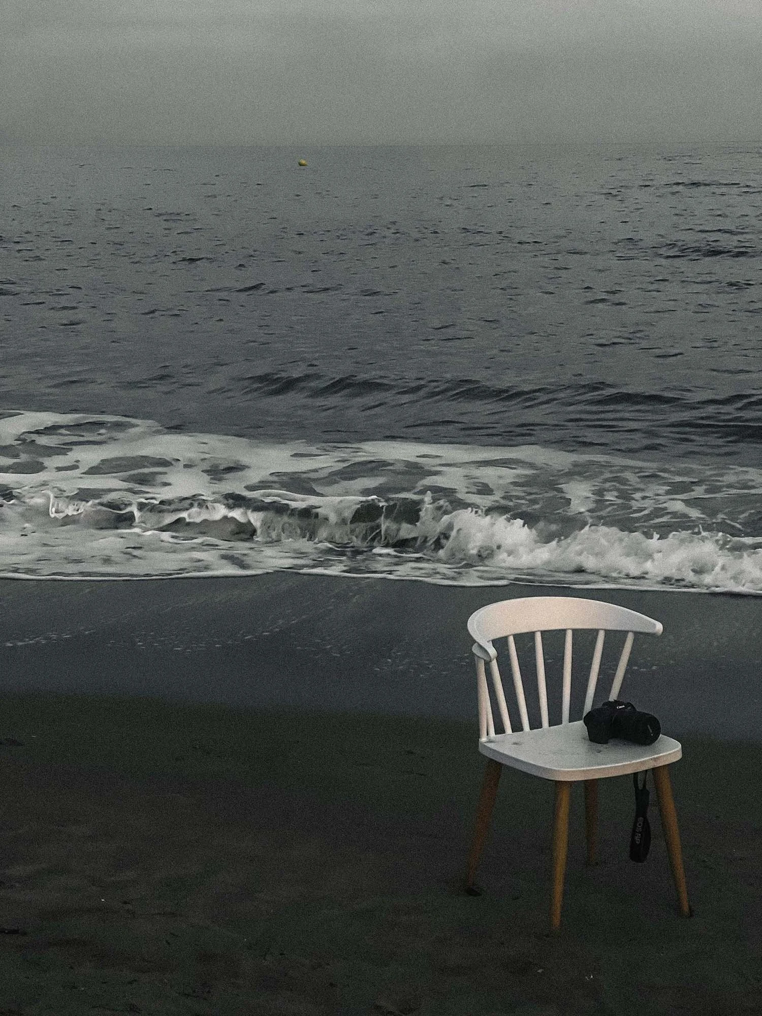 Une chaise blanche en bois avec un appareil photo posé dessus, sur une plage avec des vagues et le ciel gris en arrière-plan.