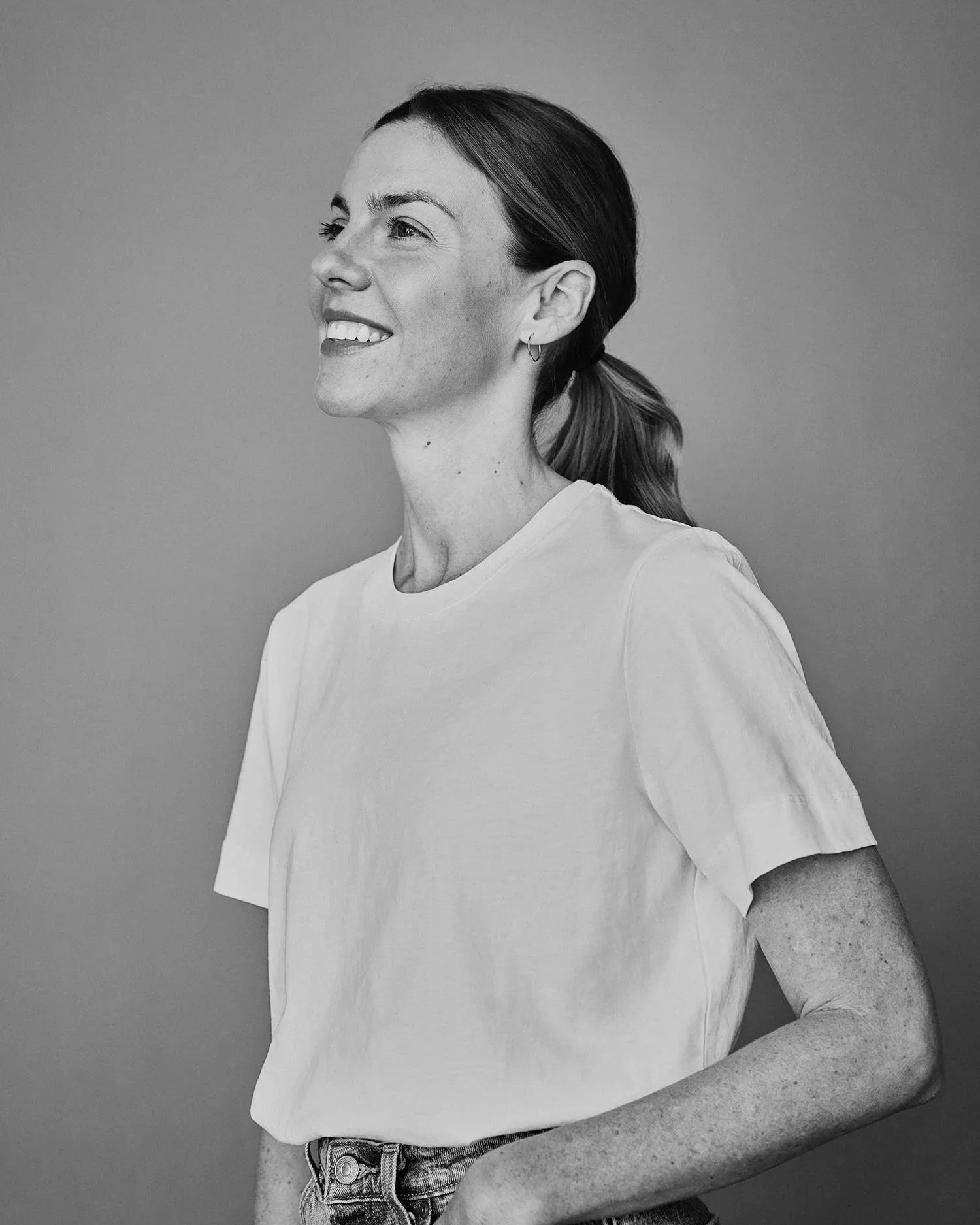 Portrait de Karina Durand, souriante, de profil, portant un t-shirt blanc et des jeans, avec des cheveux attachés en queue de cheval, en noir et blanc.