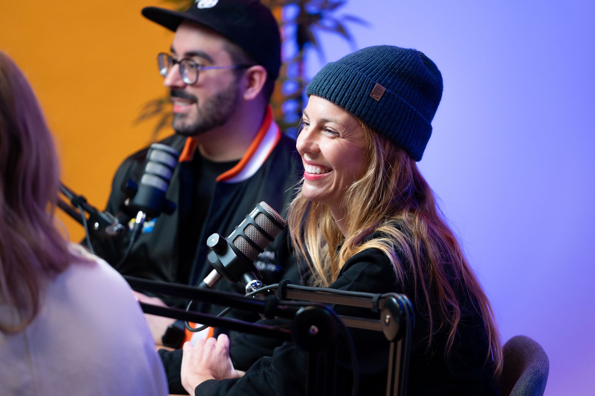 Deux personnes dont Karina Durand parlent dans un studio d'enregistrement ou un podcast, une femme avec un bonnet bleu et un homme avec des lunettes et une casquette, tous deux près de microphones.