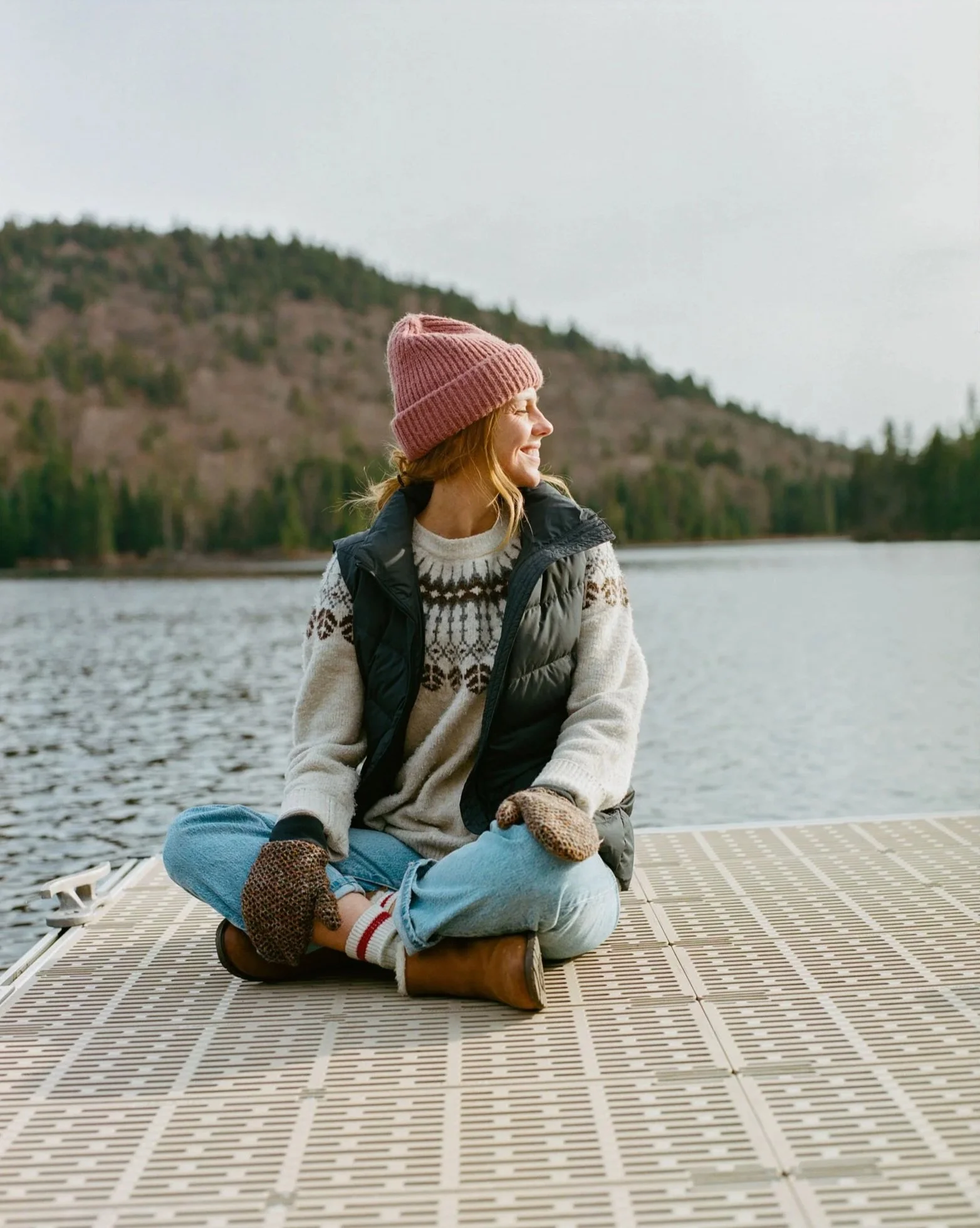 Karina Durand est assise sur une plateforme flottante au bord d'un lac, portant une écharpe, un pull à motifs, un gilet noir, un bonnet rose, des gants marron et des bottines en cuir, avec un paysage de forêt et de montagnes en arrière-plan.