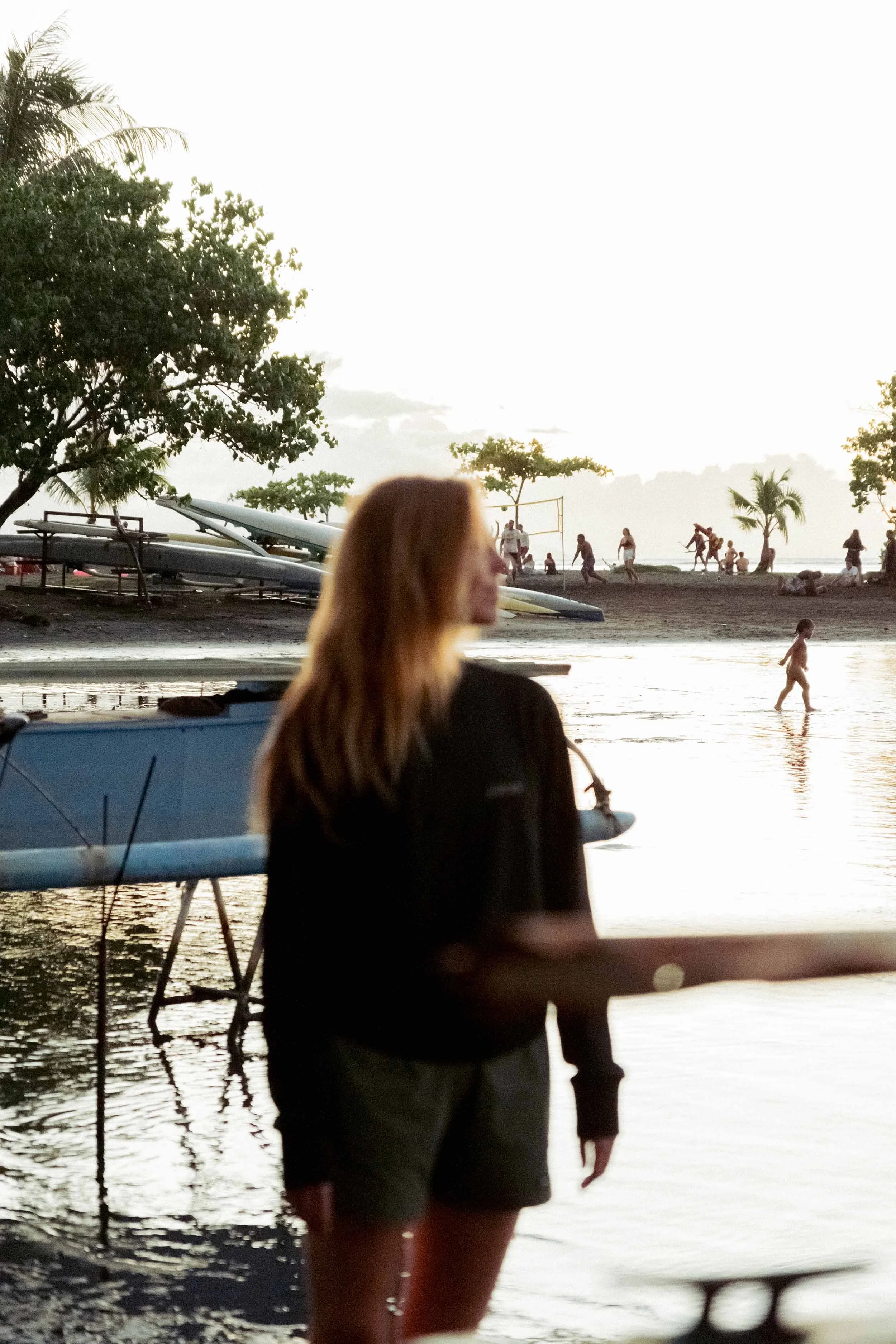 Portrait de Karina Durand portant un drysuit, face à une plage avec des kayaks, des personnes marchant, jouant et nageant dans l'eau au coucher du soleil.
