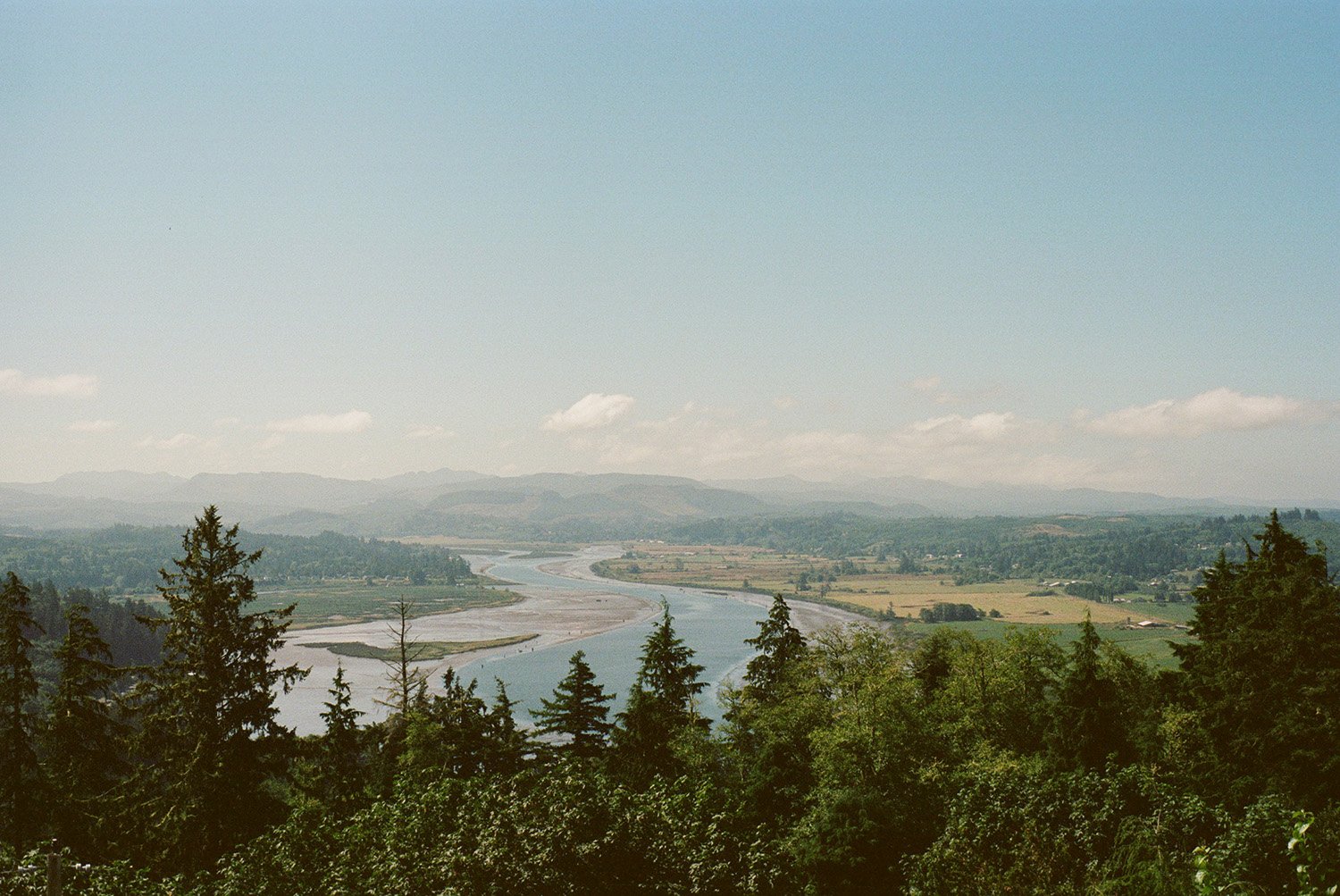 Vallée avec une rivière serpentant à travers des terrains agricoles, entourée de forêts et de collines sous un ciel clair avec quelques nuages.