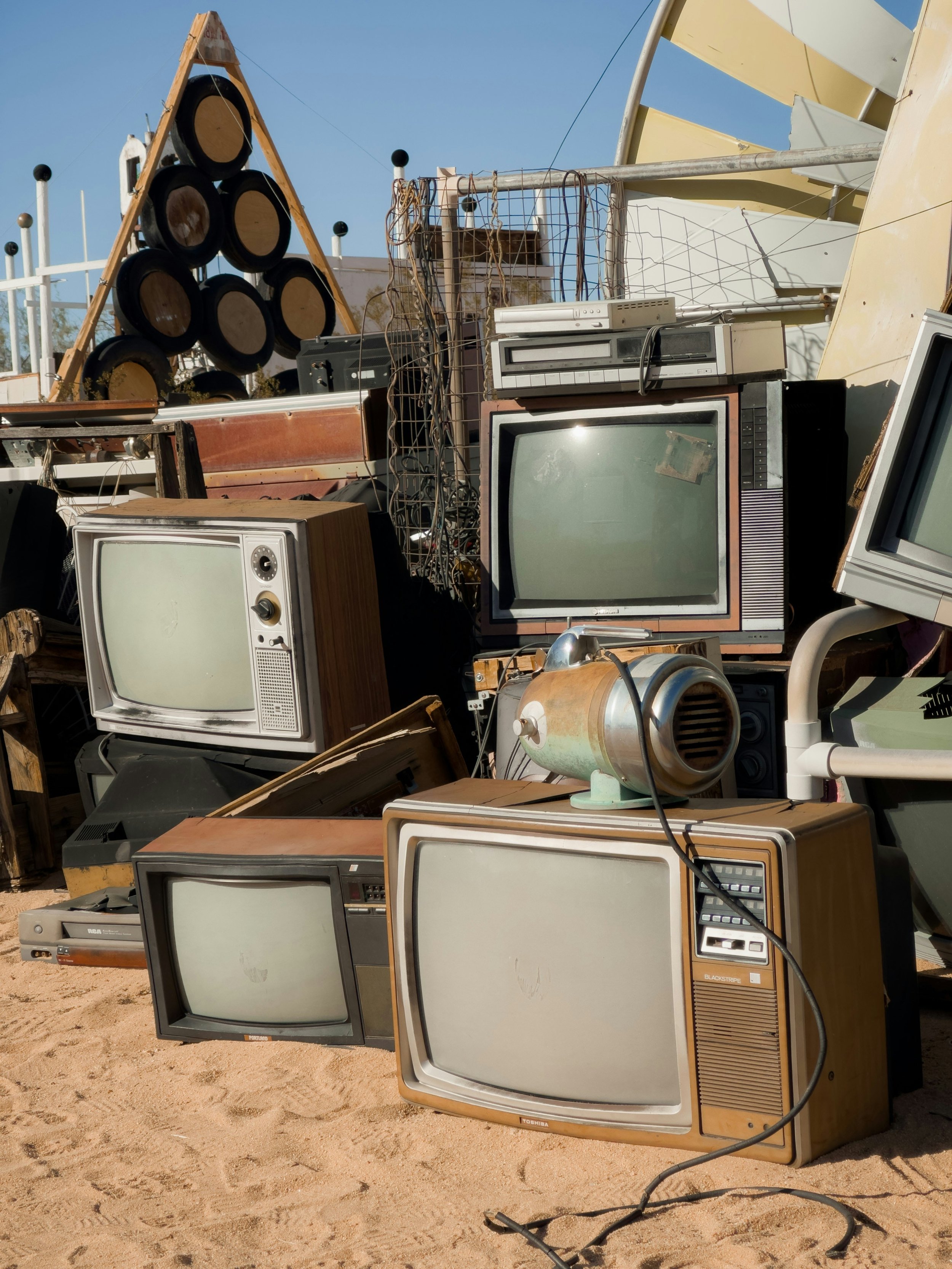 Pile of vintage and old televisions and electronic devices on sandy ground at a junkyard, with a structure of tires and a yellow building in the background.