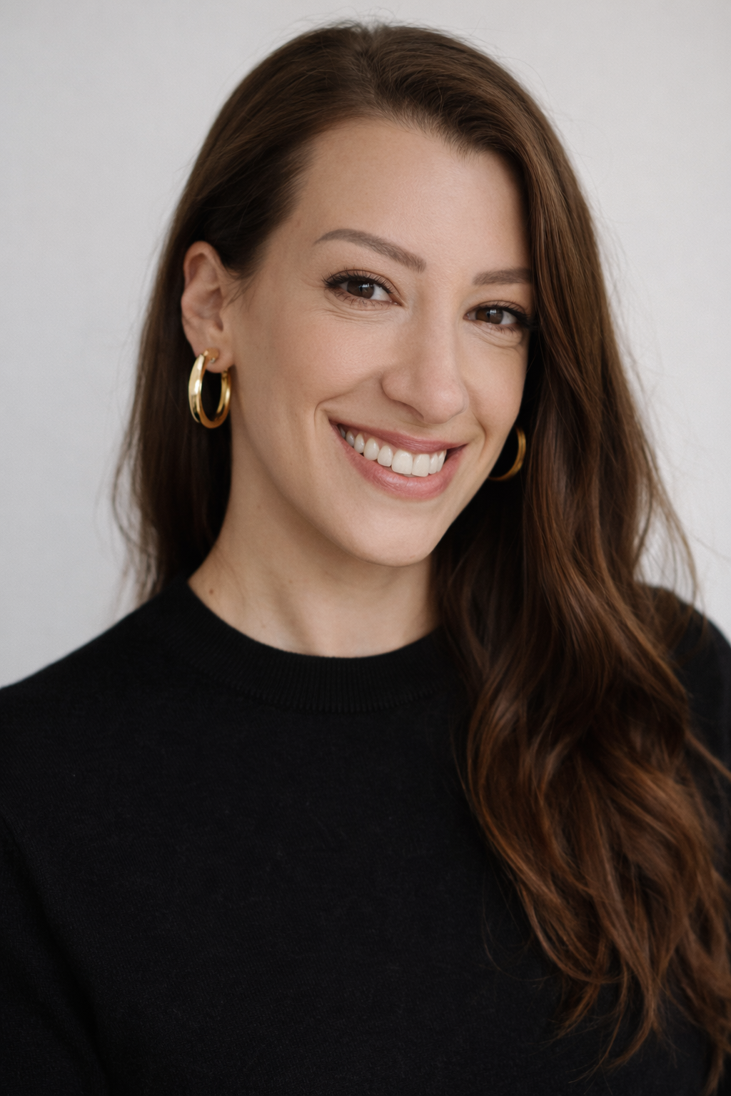 A woman with long, wavy brown hair, wearing a black top and gold hoop earrings, smiling at the camera against a plain background.
