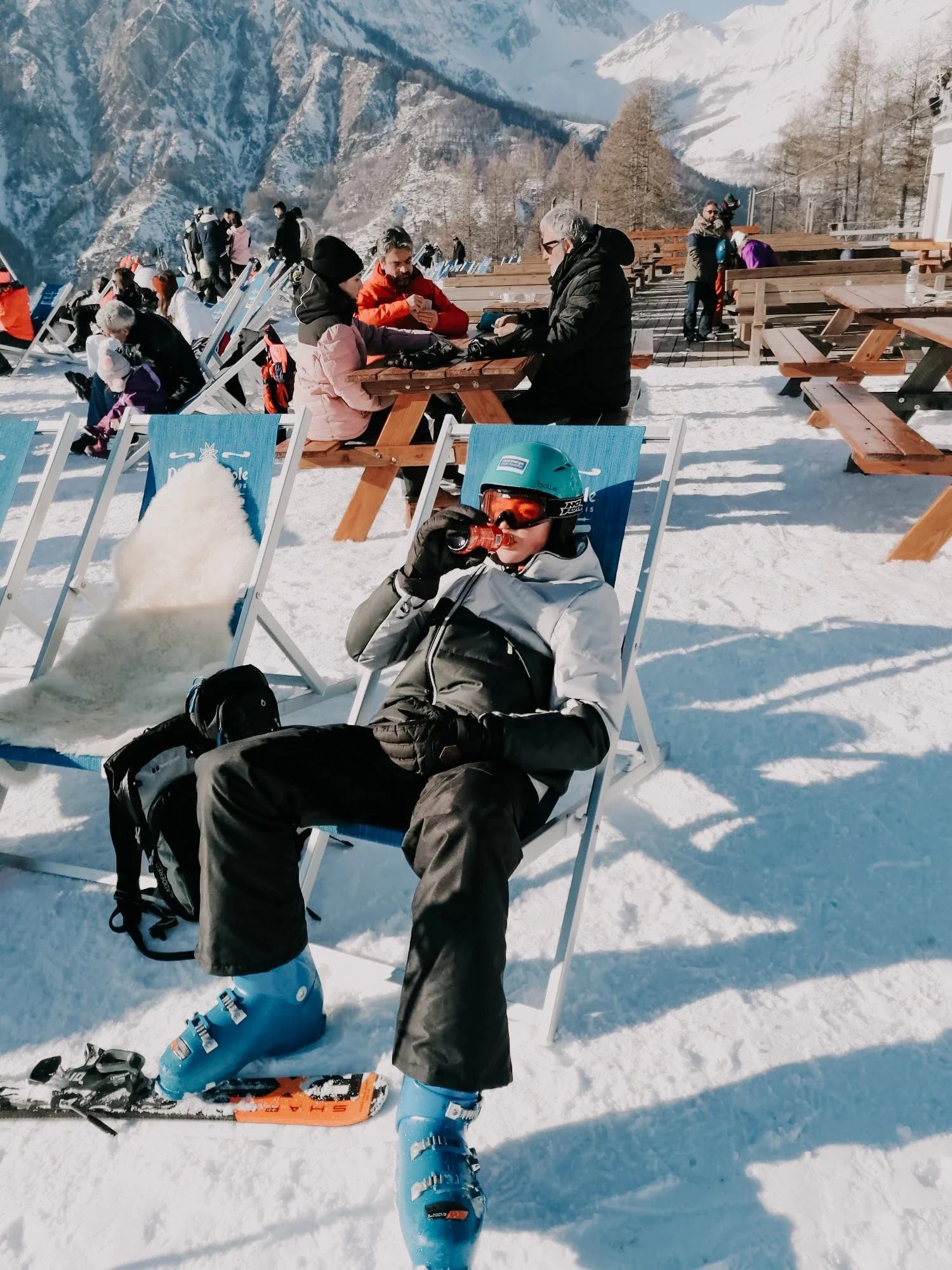 A group of people enjoying a sunny day at a snow-covered outdoor seating area with mountains in the background. Some are sitting at picnic tables, while others are standing or walking around. A child in ski gear and a helmet is seated in the foreground, drinking from a bottle.
