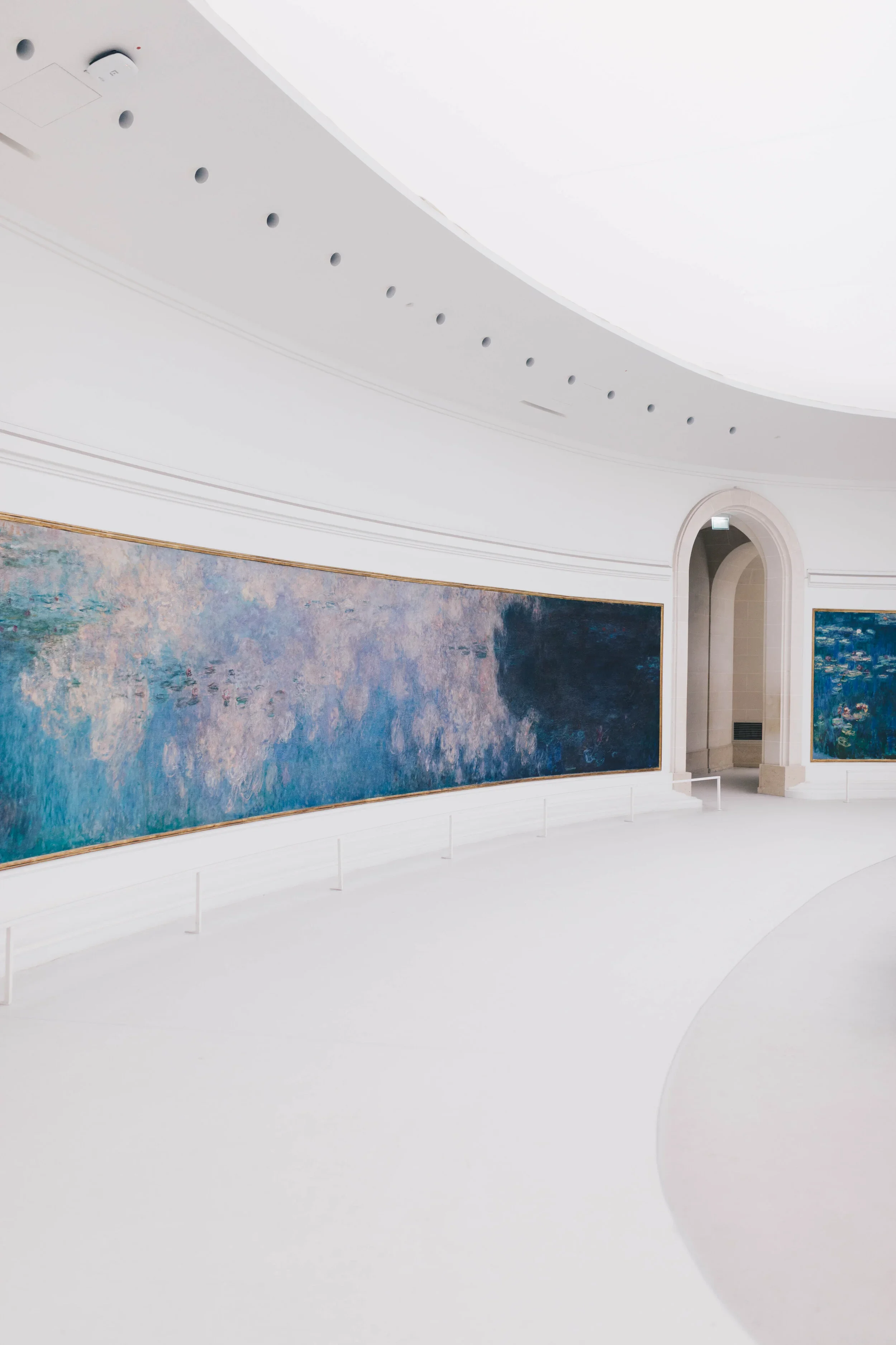 Interior view of a modern art museum gallery, showing a curved white wall with two large, abstract paintings and a high, arched doorway.