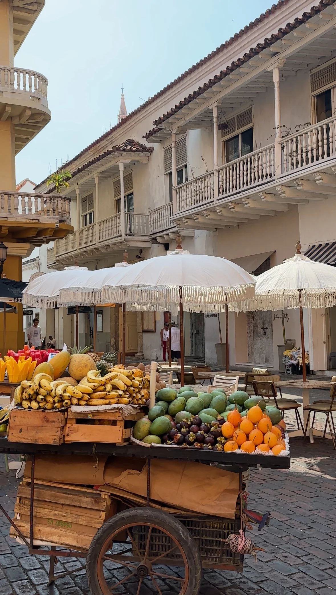 Street market scene with fresh fruits including bananas, watermelons, papayas, and oranges on wooden carts shaded by white umbrellas. The background features European-style buildings with balconies and a few people walking.