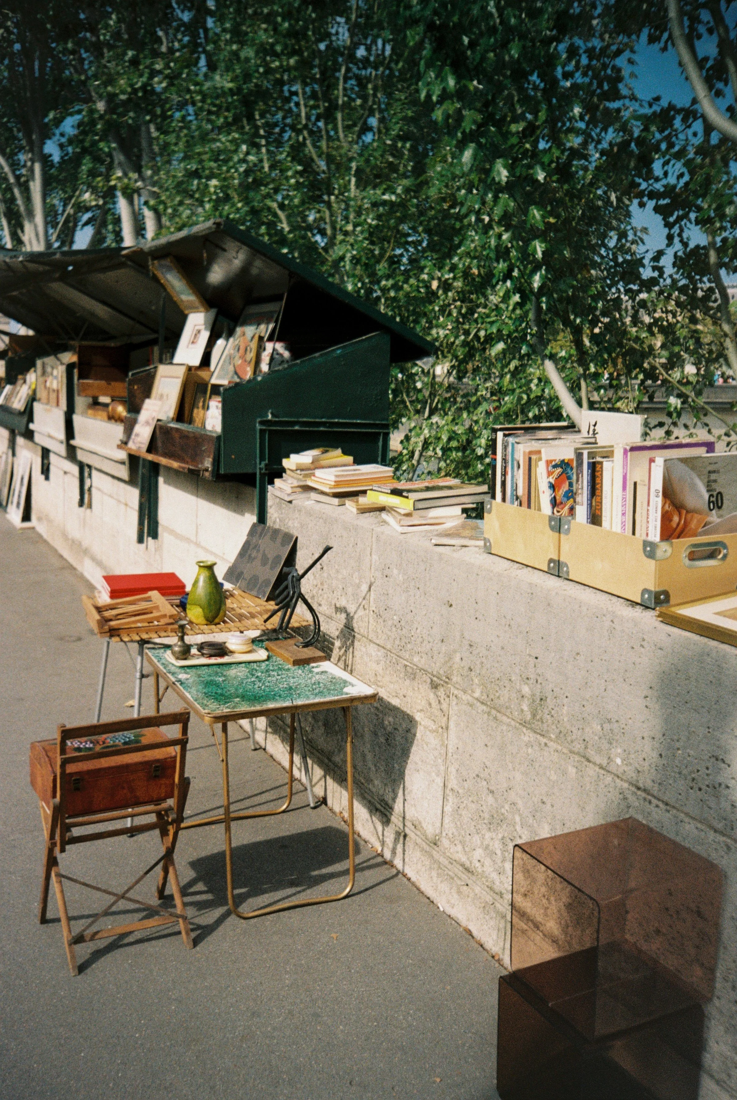 Outdoor street market or book sale with tables and boxes filled with books, paintings, and objects, set against a concrete wall and green trees under a blue sky.