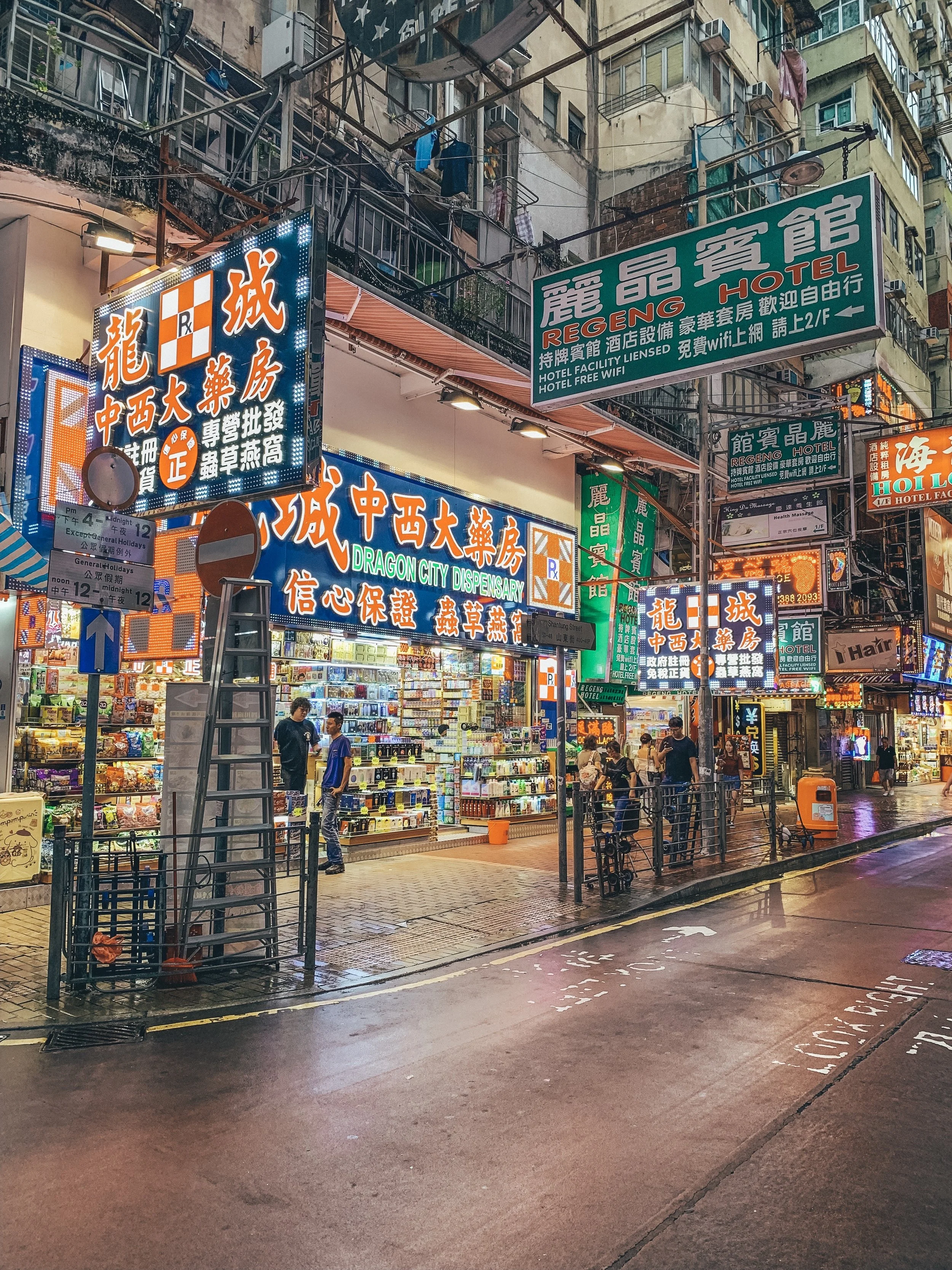 Nighttime street scene with brightly lit signs in Chinese characters, a pharmacy, a hotel named 'ReGeng Hotel,' and people walking near shops. The street is wet, reflecting the lights.