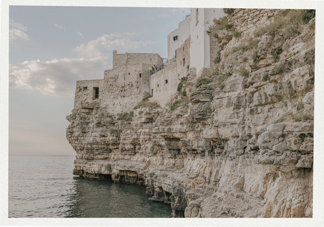 Cliffside buildings overlooking the ocean with a person relaxing on rocks below.