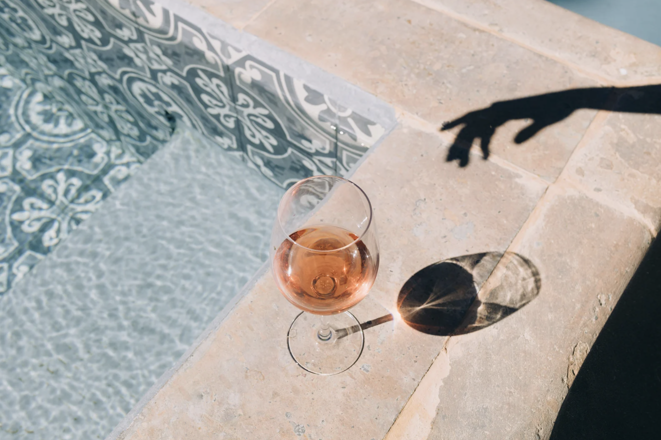 A glass of rosé wine on a stone surface next to a pool. The shadow of a curved metal pool ladder is cast on the surface.