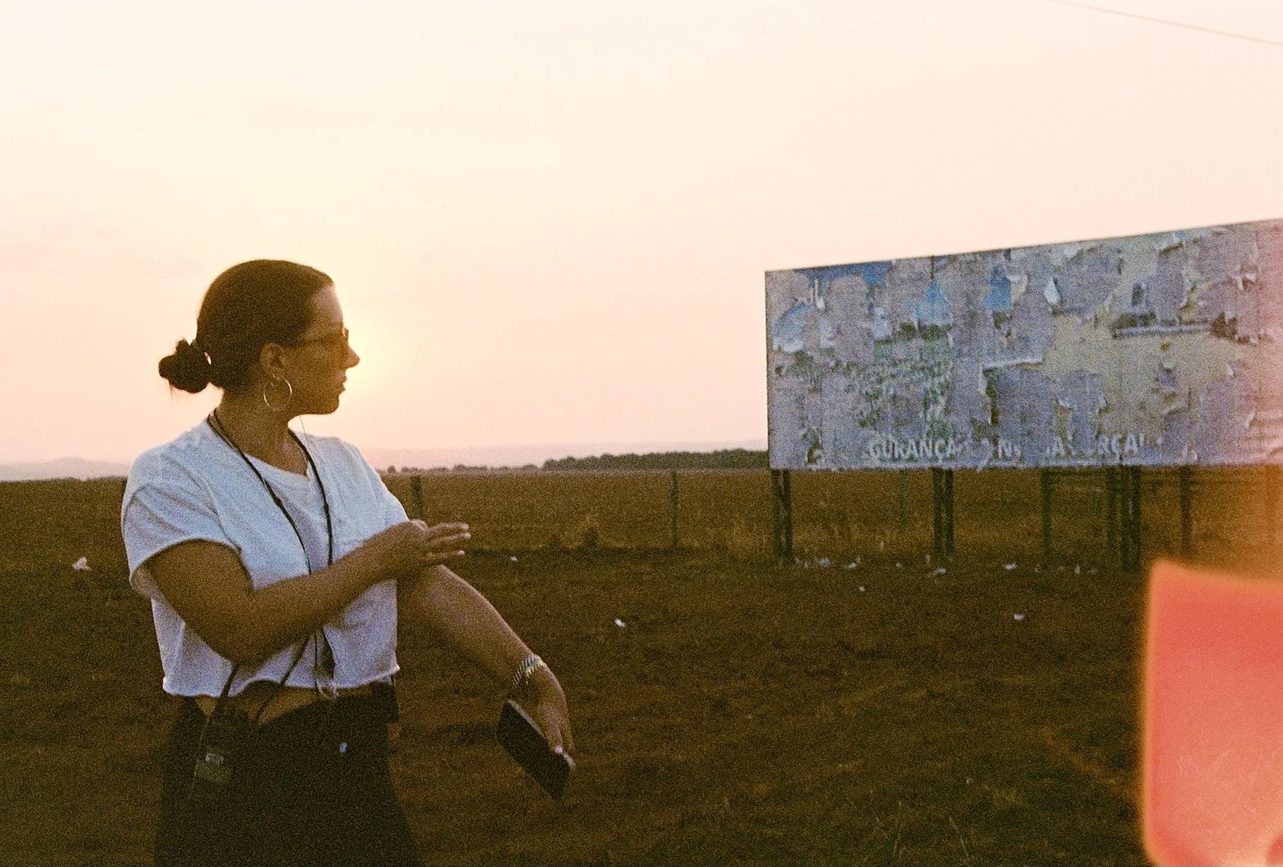 A woman standing on a dry field during sunset, with her arms crossed, holding a phone, and looking towards an old, worn billboard in the background.