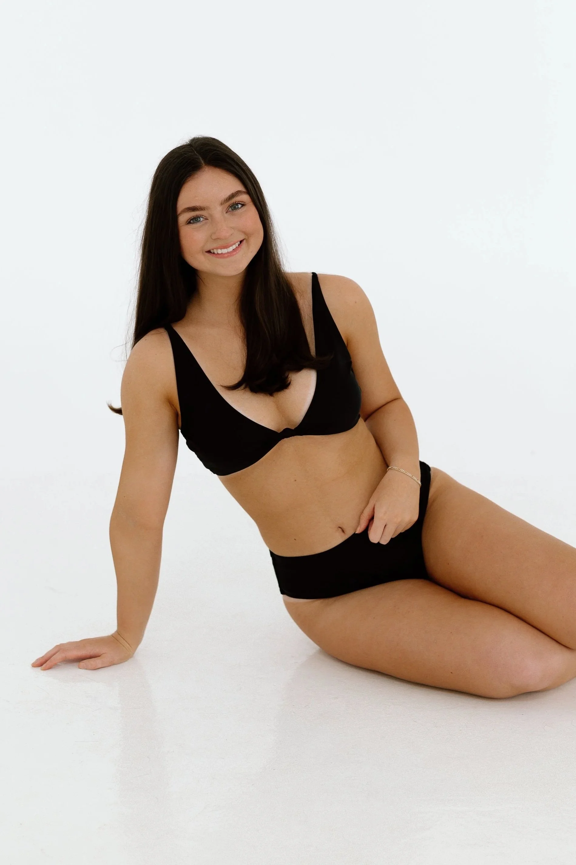 Young woman with long dark hair sitting on the floor in black lingerie, smiling at the camera against a plain white background.