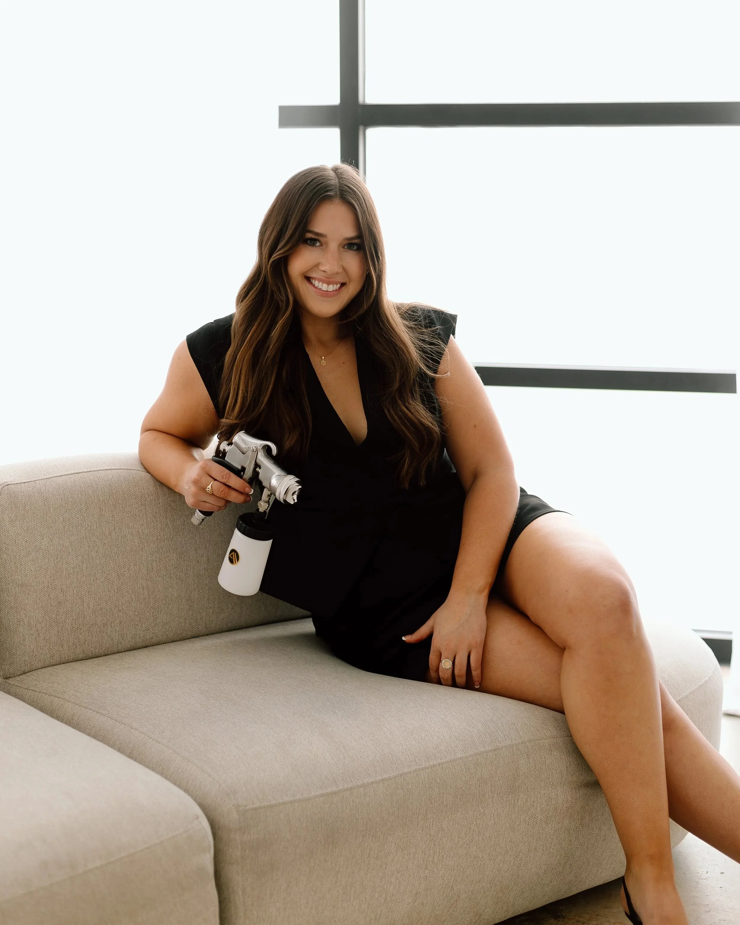 A woman with long brown hair sitting on a beige sofa near a large window, smiling, holding a telescope, wearing a black dress and jewelry.