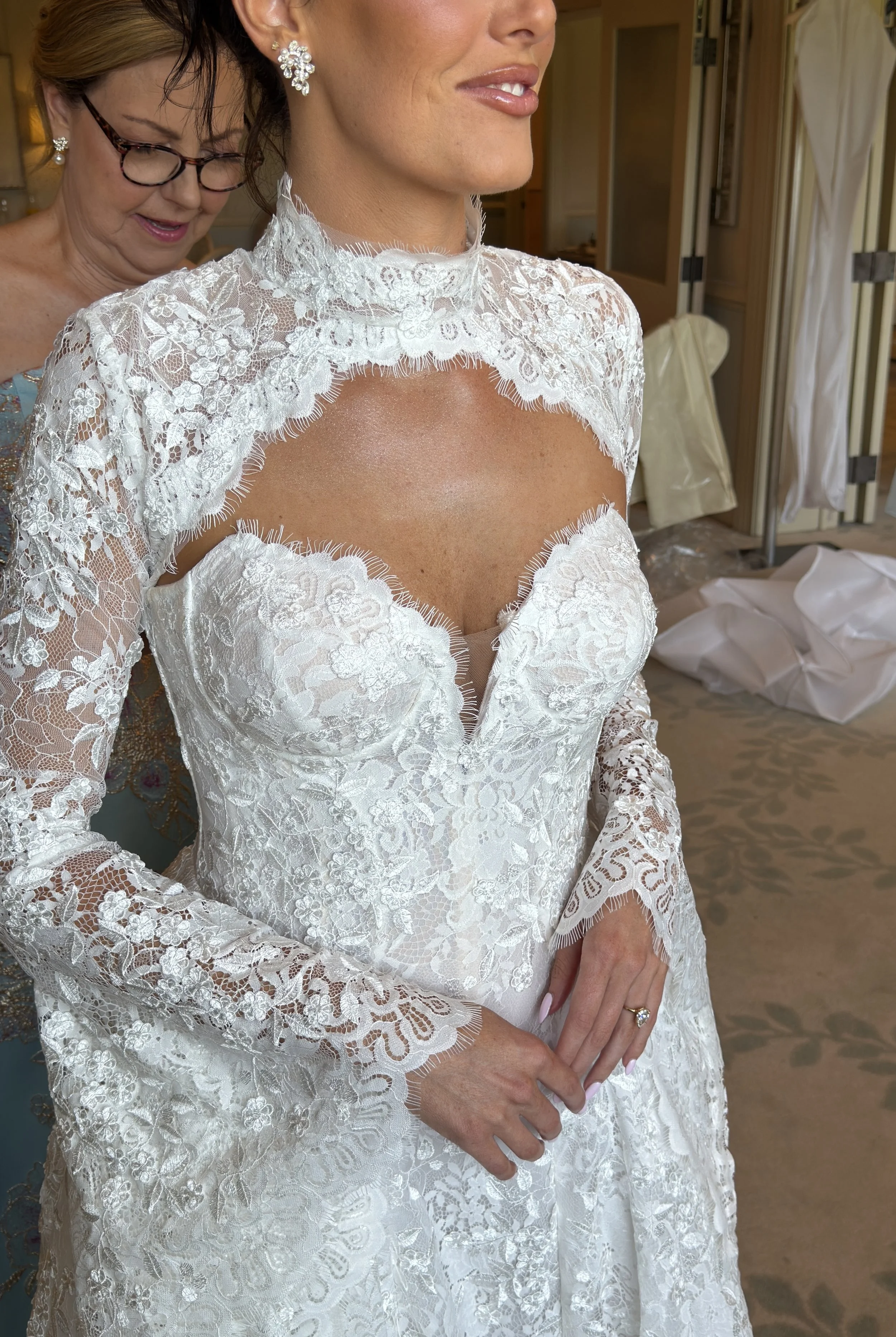 Close-up of a woman in a white lace wedding dress with floral patterns, wearing pearl earrings and a ring, with an older woman helping her get ready.