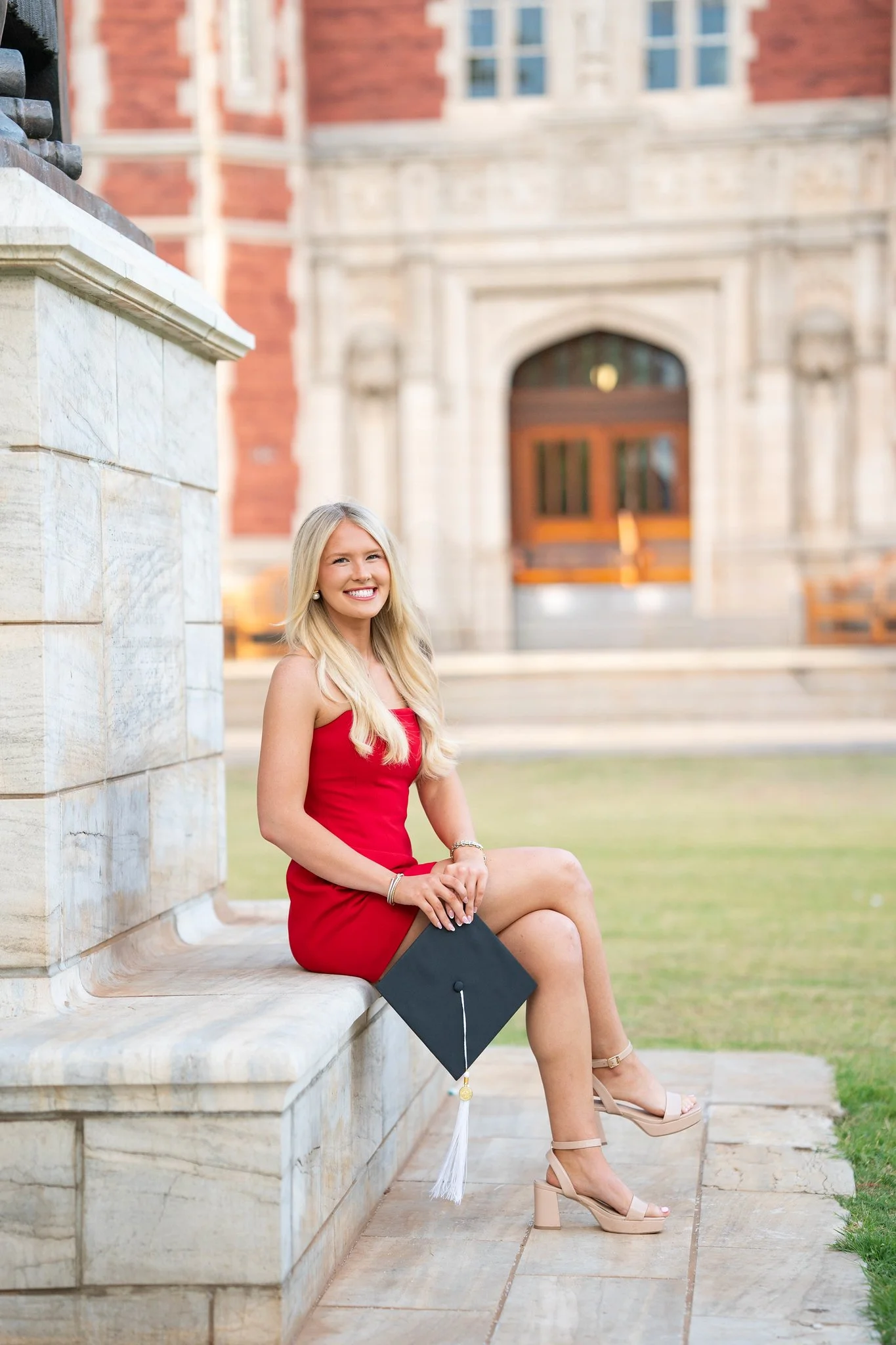 A young woman with long blonde hair wearing a red dress and beige platform heels sitting on a marble ledge outside a historic brick building, holding a black graduation cap with a white tassel.
