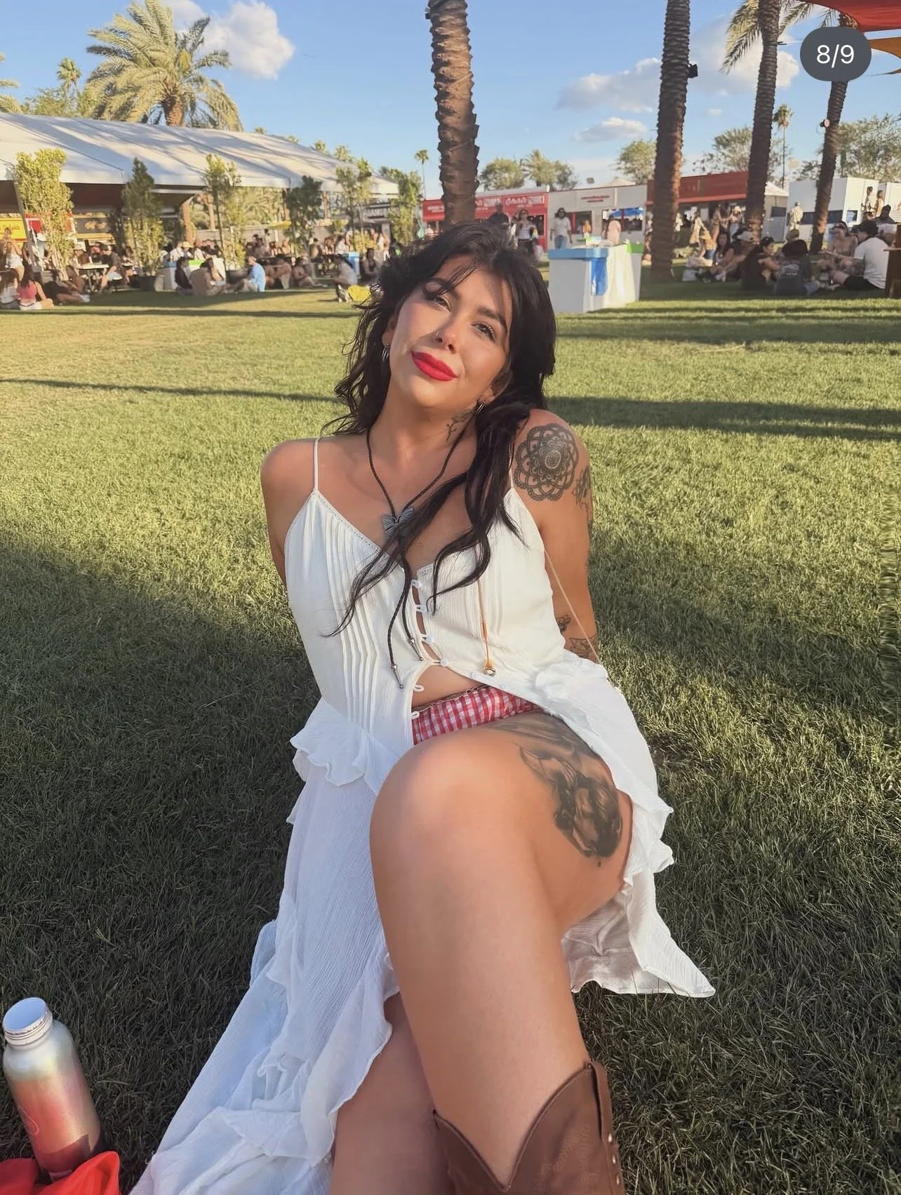A woman with dark hair, red lipstick, and tattoos sitting on the grass at an outdoor event with palm trees, tents, and many people in the background during the daytime.