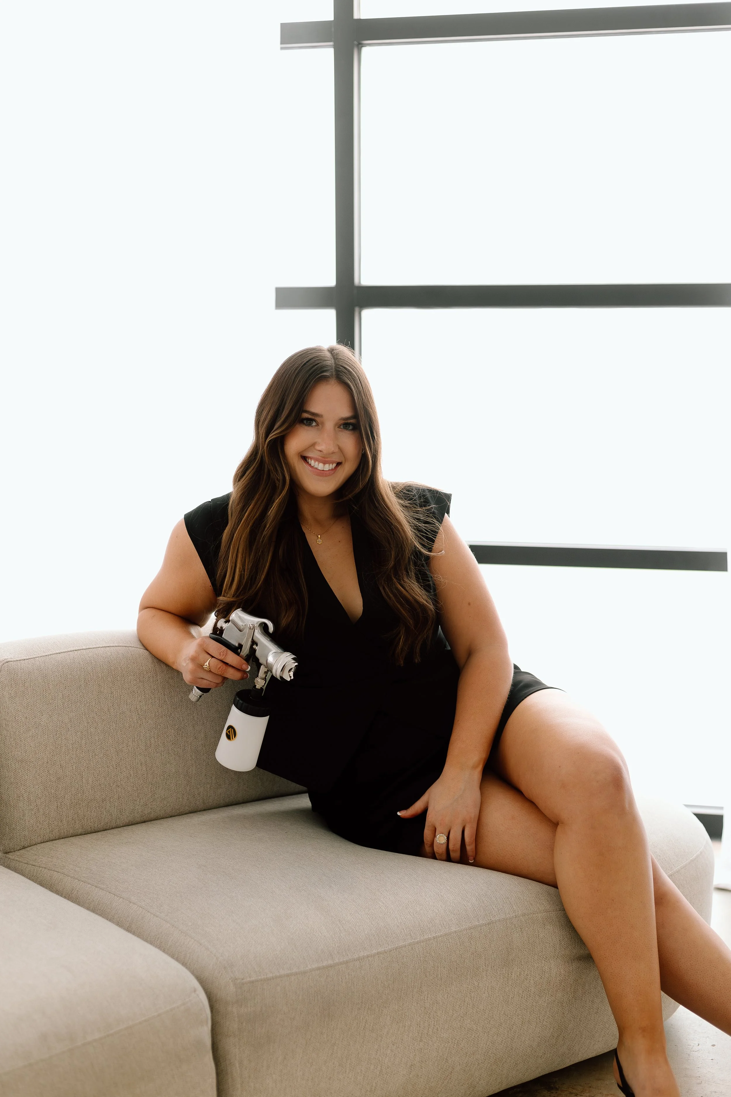 A woman with long brown hair, smiling, sitting on a beige sofa, holding a spray gun, wearing a black dress, and black slippers, in a room with large window panels.