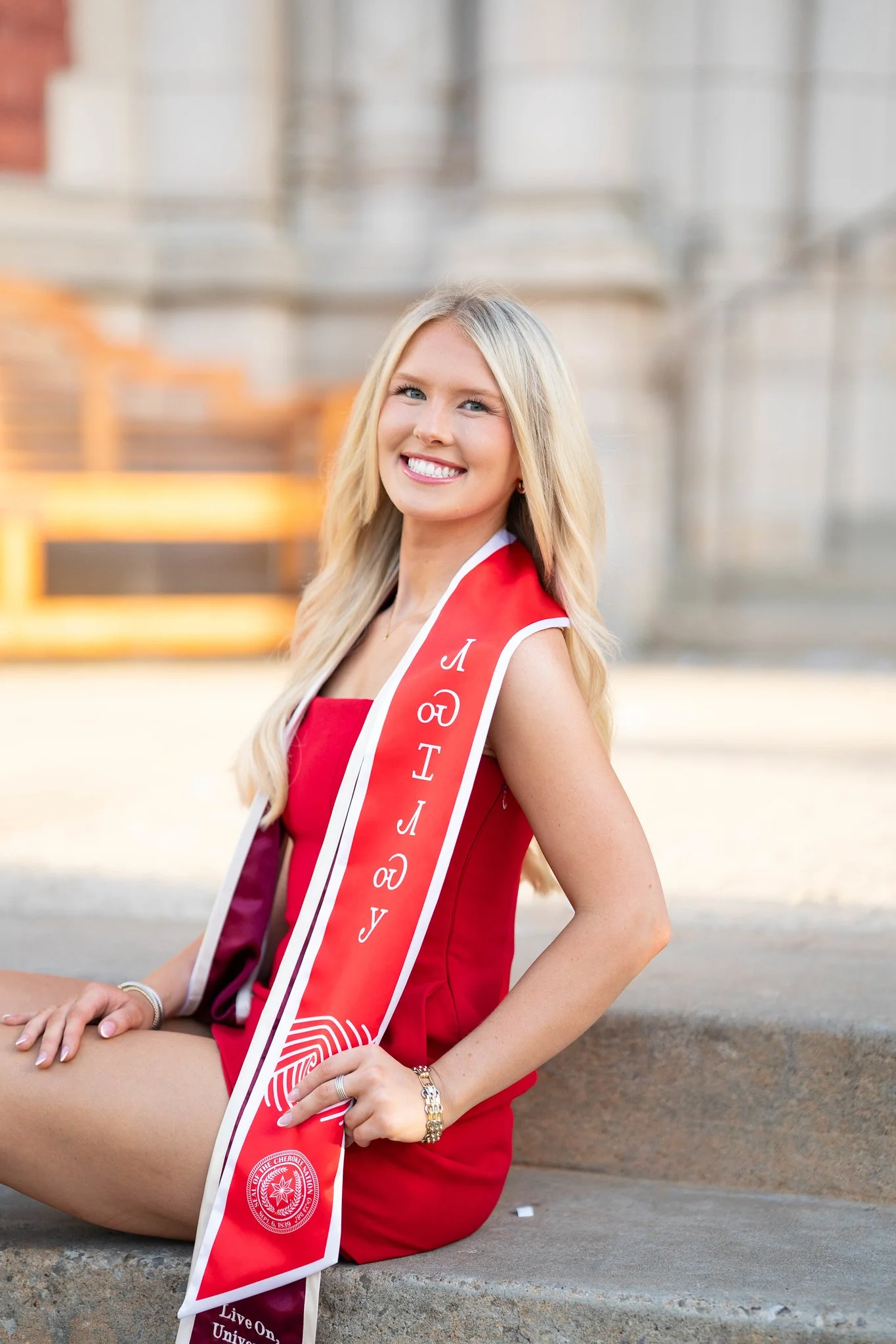 A young woman with long blonde hair, wearing a red dress and a red sash with white trim and lettering, sitting on a stone curb in front of a historical building with columns. She is smiling and posing for a photo.